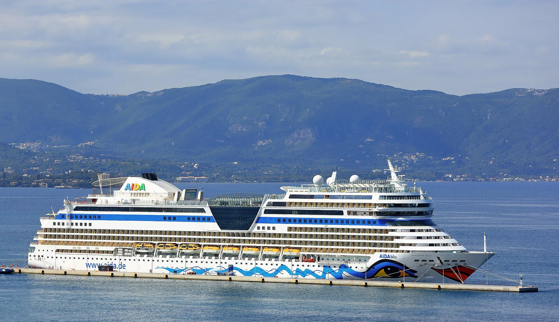 Das Kreuzfahrtschiff AIDAblu der Sphinx-Klasse am Kai im Hafen von Korfu vor bewaldeten Bergen.