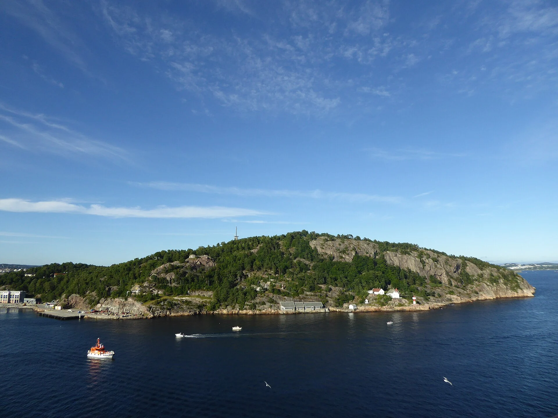 Die bewaldete Felsenhalbinsel Odderøya bei Kristiansand vom Wasser aus gesehen, mit Leuchtturmstation und alten Militärgebäuden am Ufer.