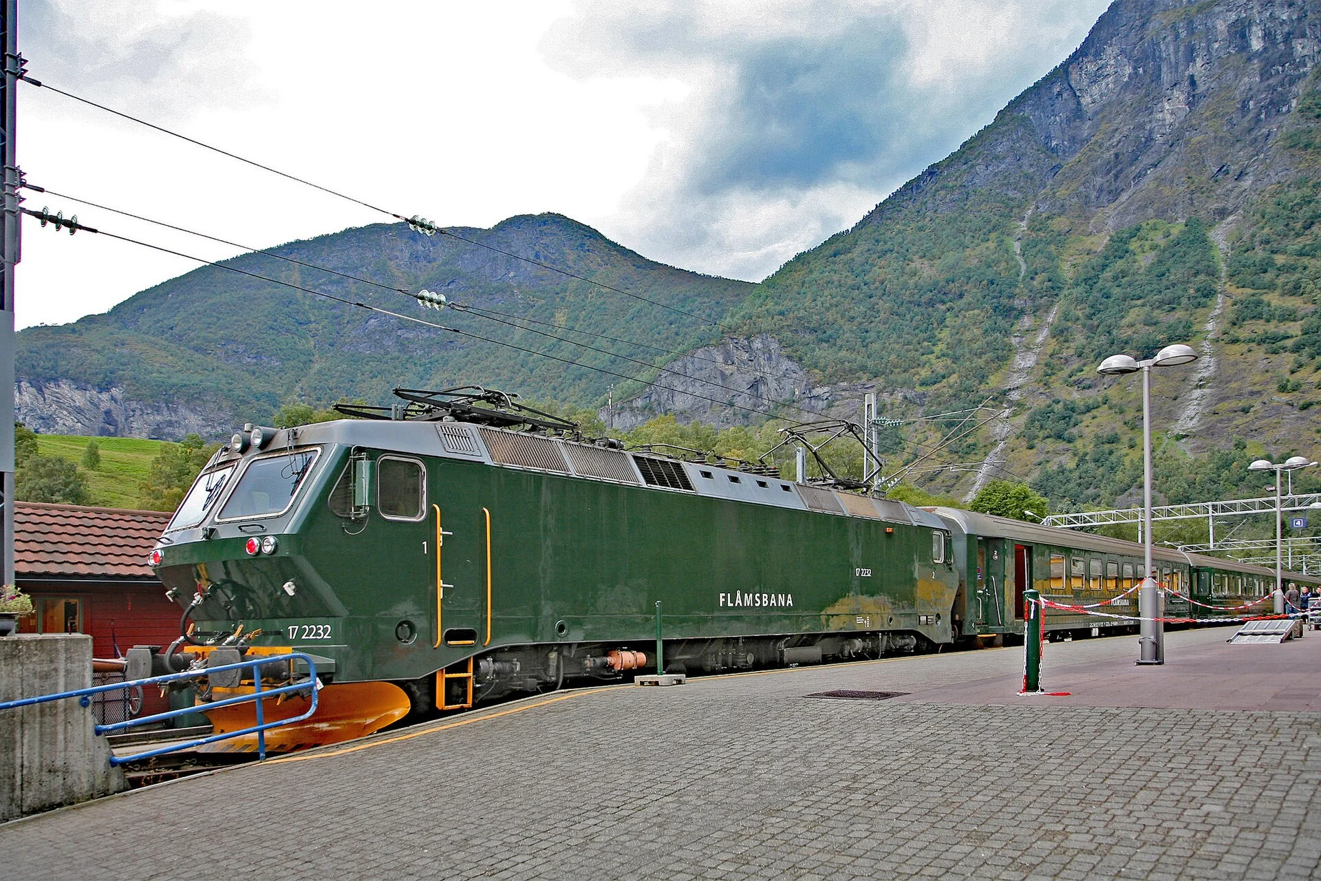 Grüne Lokomotive mit Aufschrift „Flåmsbana“ mit Zug am Bahnhof Flåm vor steilen Berghängen, Norwegen.