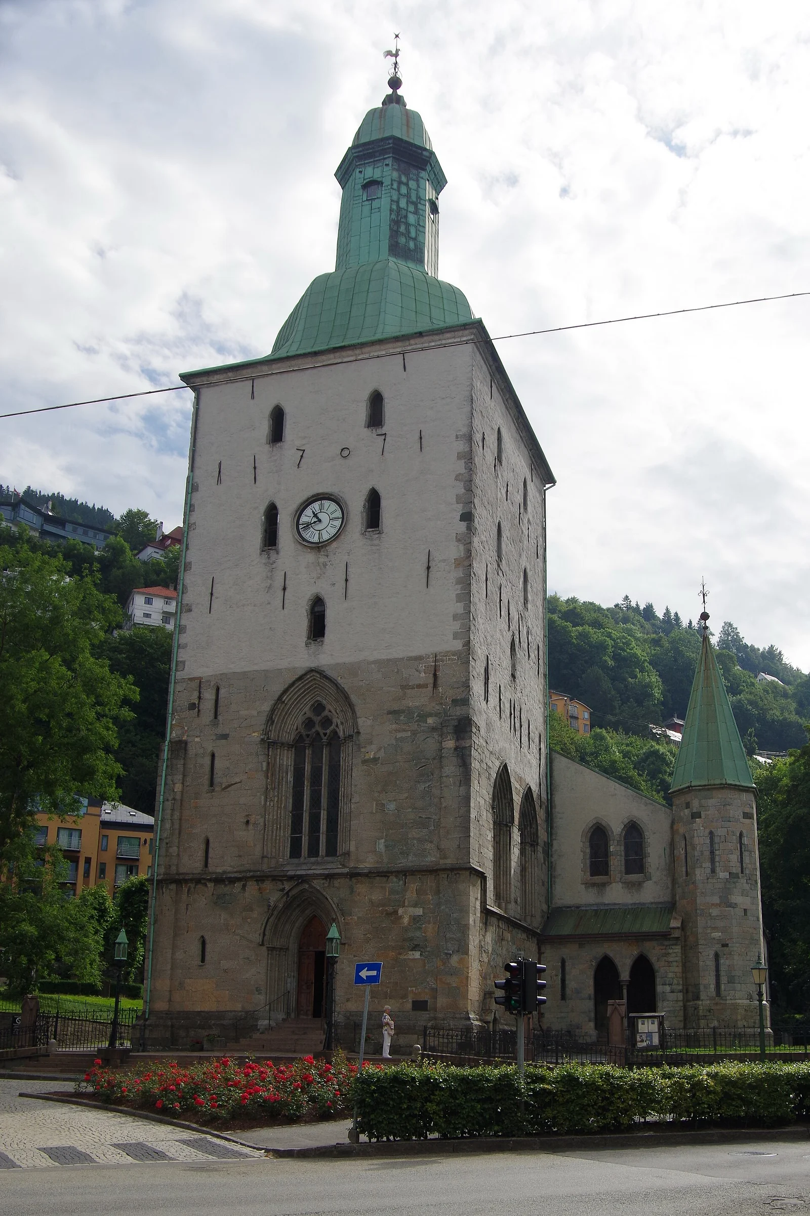 Turmfassade und Südseite der Bergen Domkirke in Bergen, Norwegen, mit Blumenbeet und Straßenkreuzung im Vordergrund.