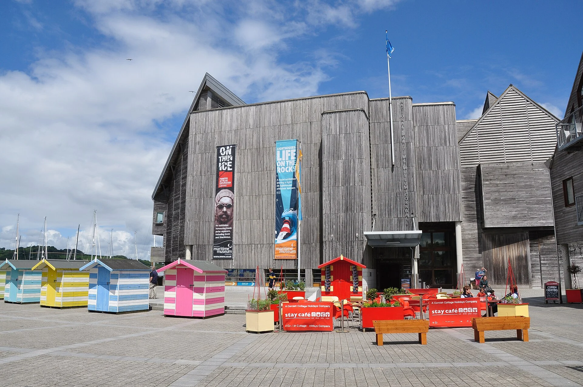 Das mit Holz verkleidete Gebäude des National Maritime Museum Cornwall am Discovery Quay in Falmouth mit bunten Strandhütten auf dem Vorplatz.