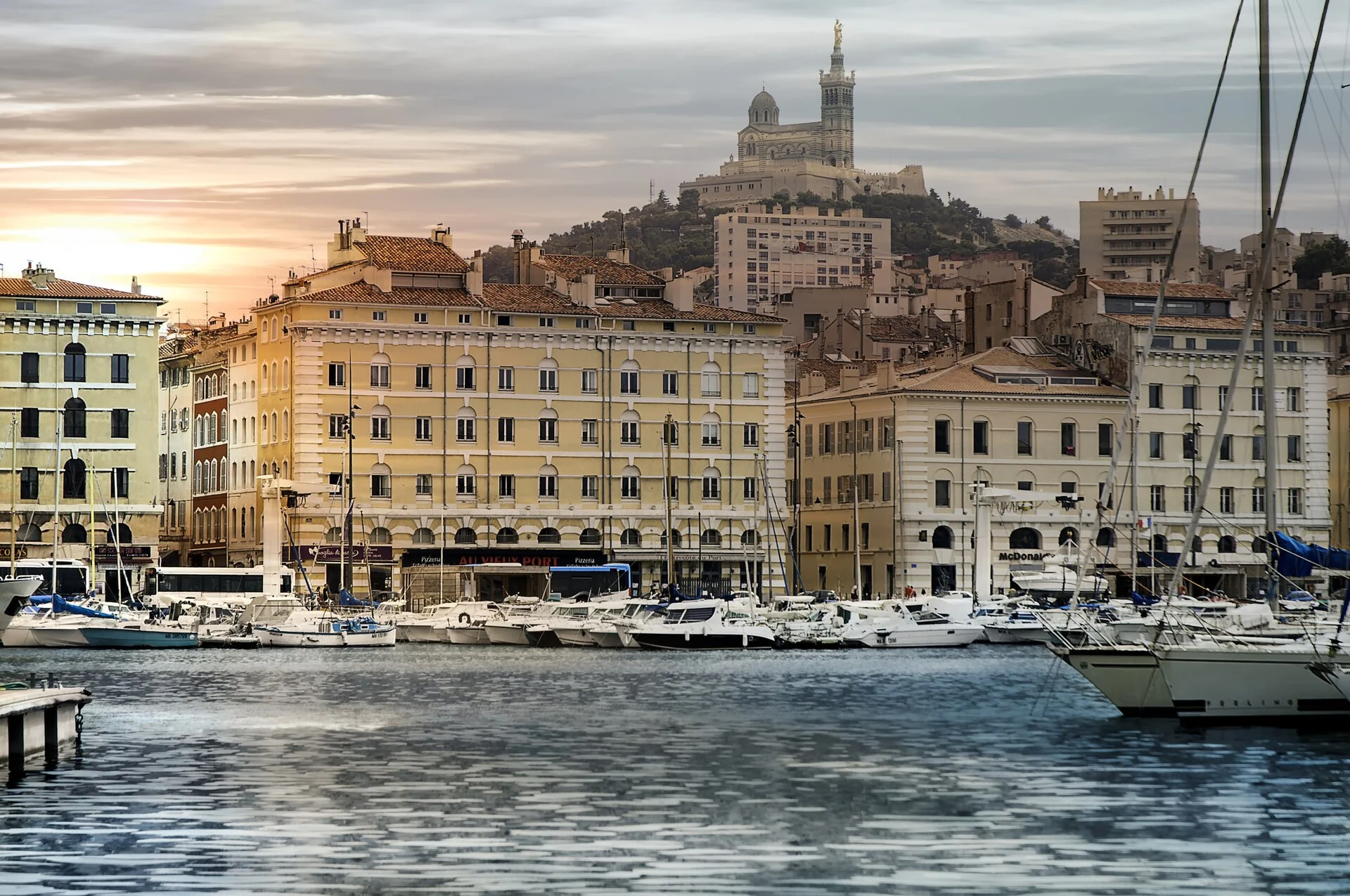 Der Vieux-Port von Marseille mit vertäuten Yachten vor den Kaifassaden und der Basilika Notre-Dame de la Garde auf dem Hügel im Hintergrund.
