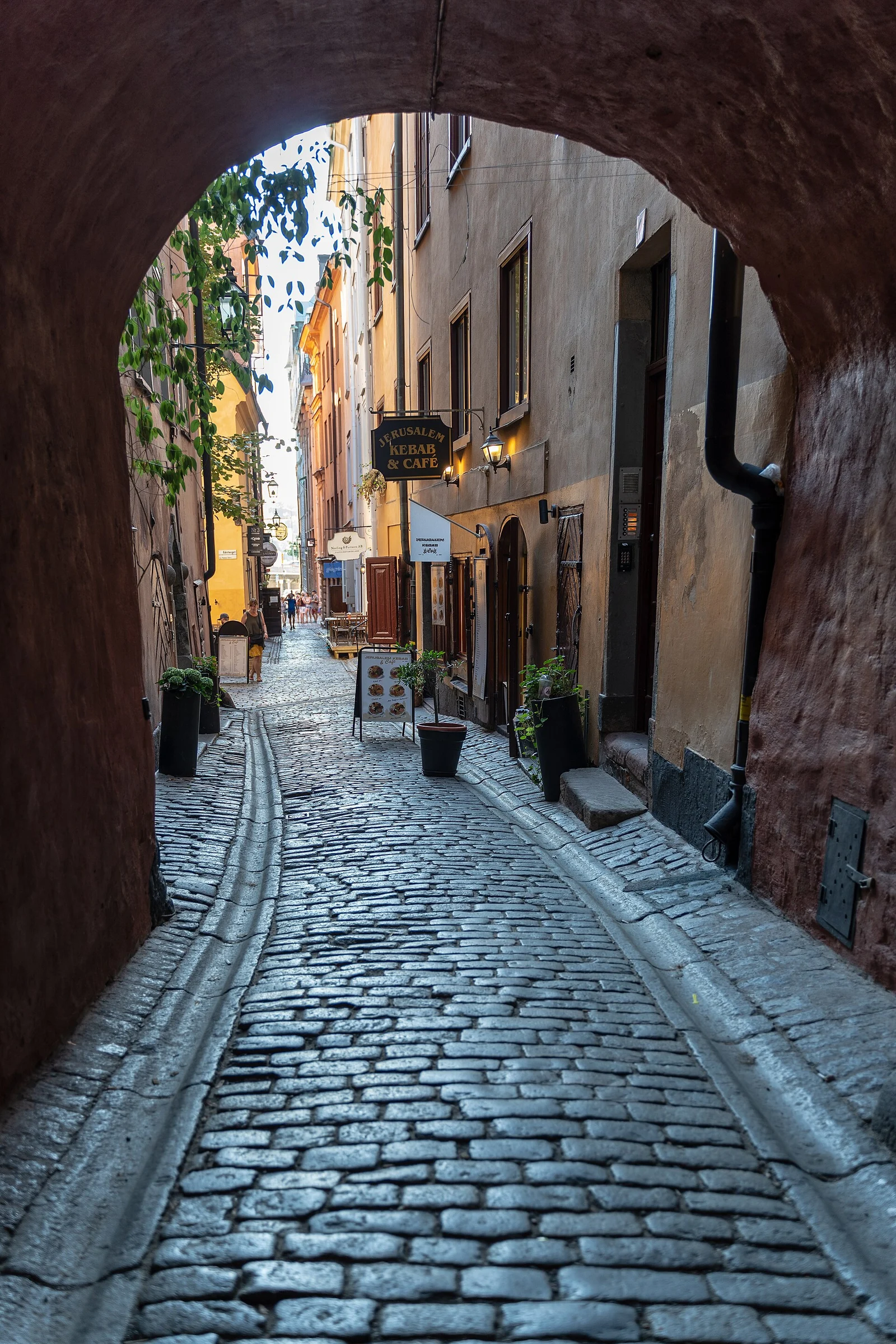 Blick durch einen Torbogen in eine enge, kopfsteingepflasterte Gasse mit Läden und Cafés in Gamla Stan, Stockholm.