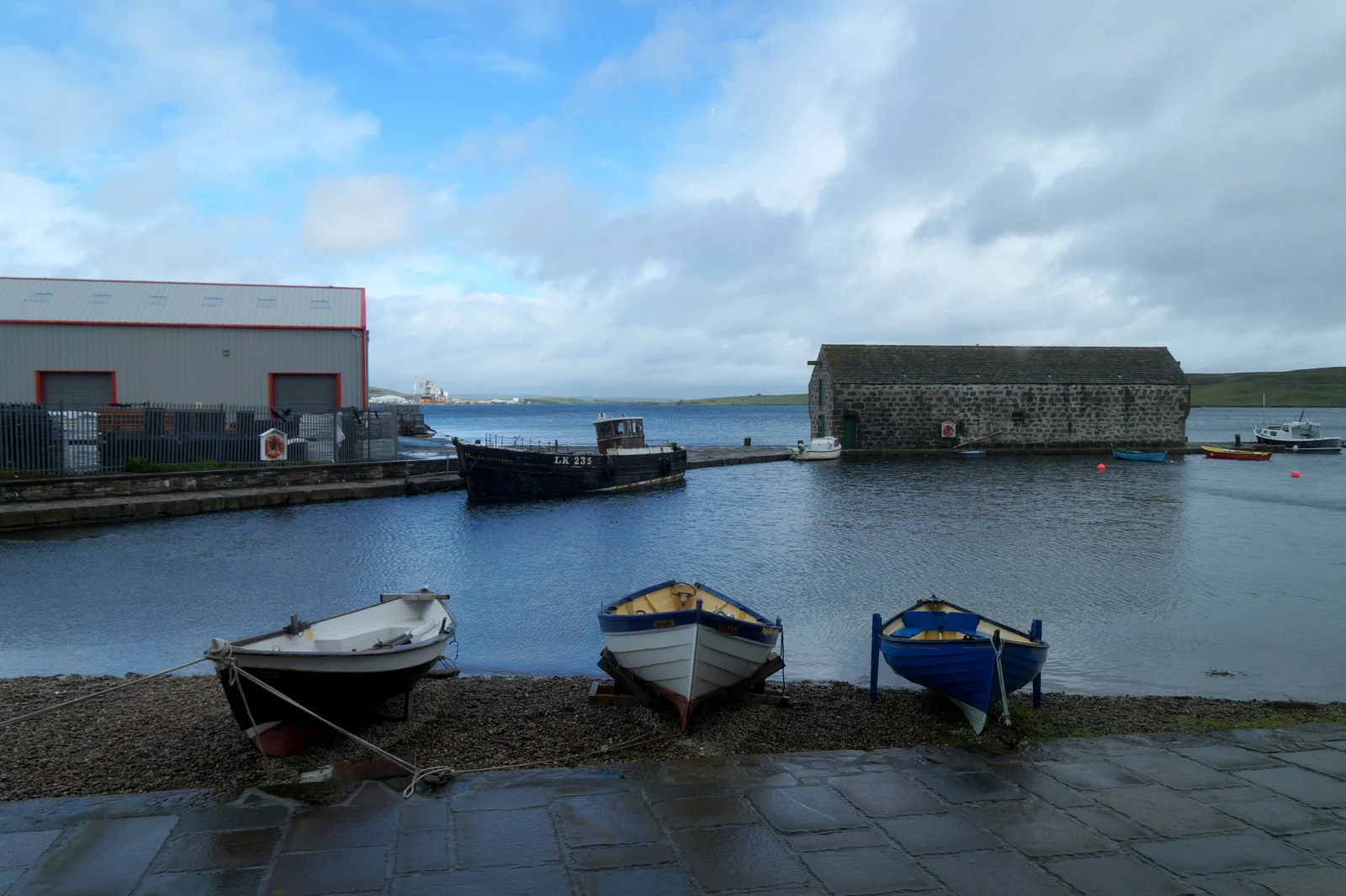 Blick vom Shetland Museum and Archives über Hay's Dock in Lerwick mit Ruderbooten am Ufer und historischem Bootshaus.