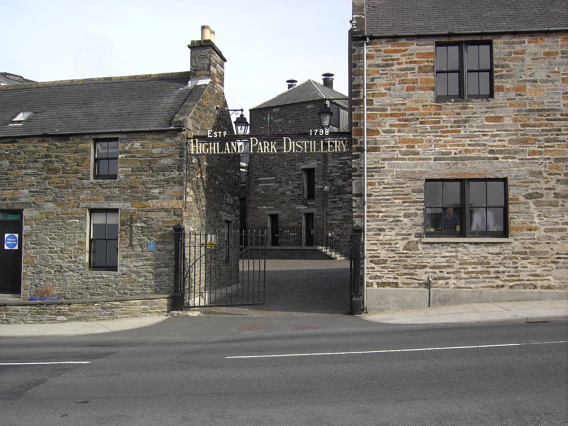 Eingangstor der Highland Park Distillery in Kirkwall mit Schriftzug „Highland Park Distillery Estd 1798“ über der Toreinfahrt zwischen Sandsteingebäuden.