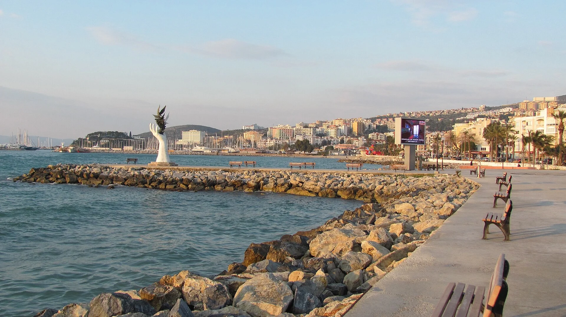 Uferpromenade von Kuşadası mit Friedensskulptur, Wellenbrecher und Stadtkulisse.