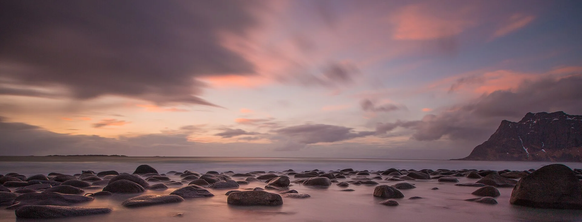 Runde Felsbrocken in der flachen Brandung am Strand von Uttakleiv bei Leknes auf den Lofoten, im Abendlicht mit rosa Wolken und steilem Berg am rechten Bildrand.