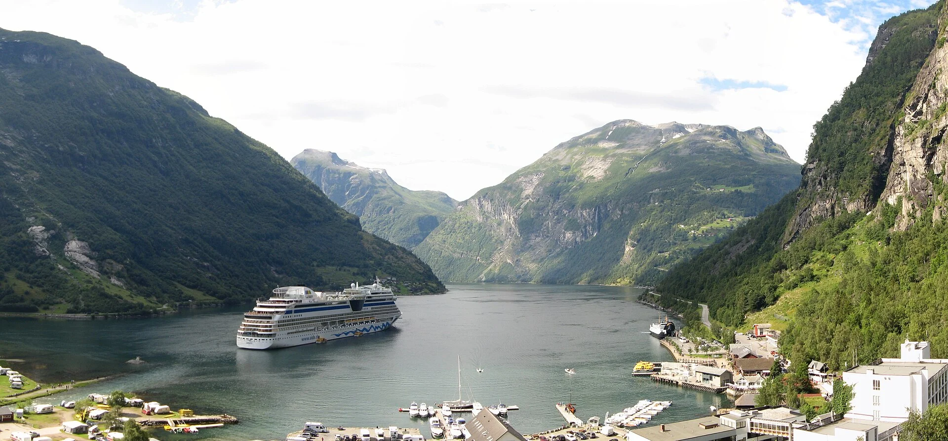 Blick über Geiranger und den Geirangerfjord mit Kreuzfahrtschiff, rechts die Hangbebauung des Ortes mit dem Bereich des Norsk Fjordsenter.