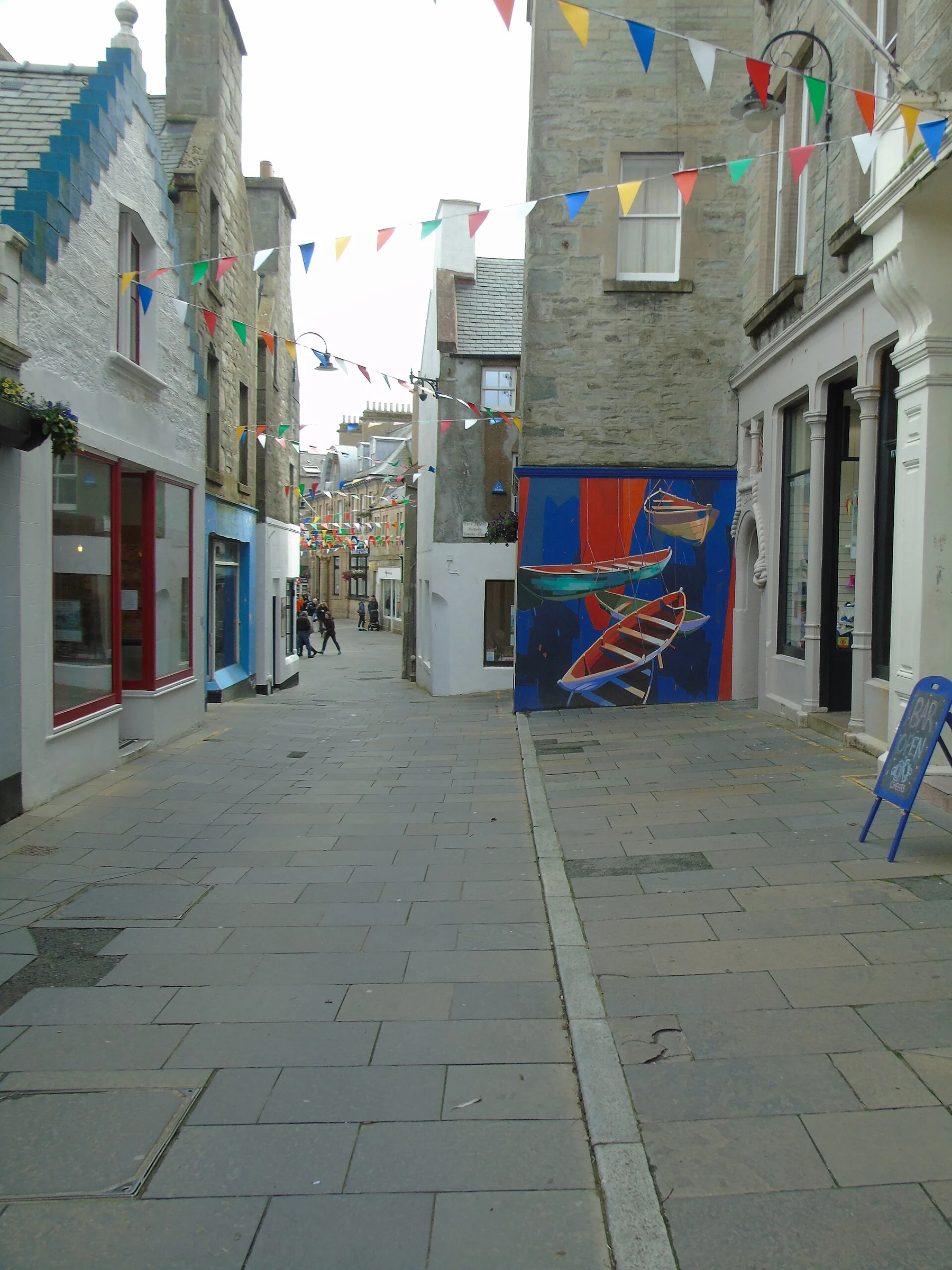 Blick in die gepflasterte Commercial Street in Lerwick mit Wimpelketten, Steinhäusern und einem Wandbild mit Ruderbooten an der Einmündung der Pitt Lane.