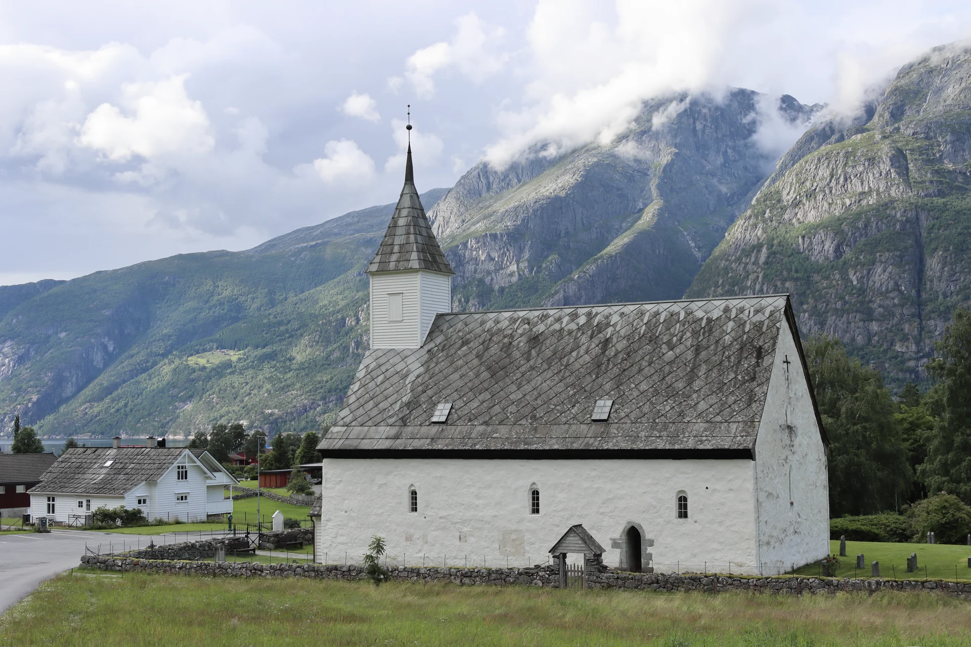 Die weiß verputzte Steinkirche Eidfjord gamle kyrkje mit Schieferdach, Dachreiter und Friedhofsmauer vor steilen Bergen in Eidfjord, Norwegen.