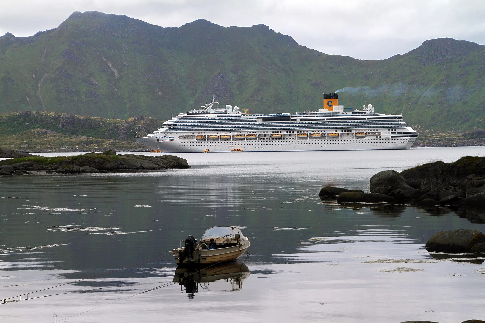 Ein Kreuzfahrtschiff liegt im Fjord bei Leknes auf den Lofoten, im Vordergrund Felsen und ein kleines Boot.
