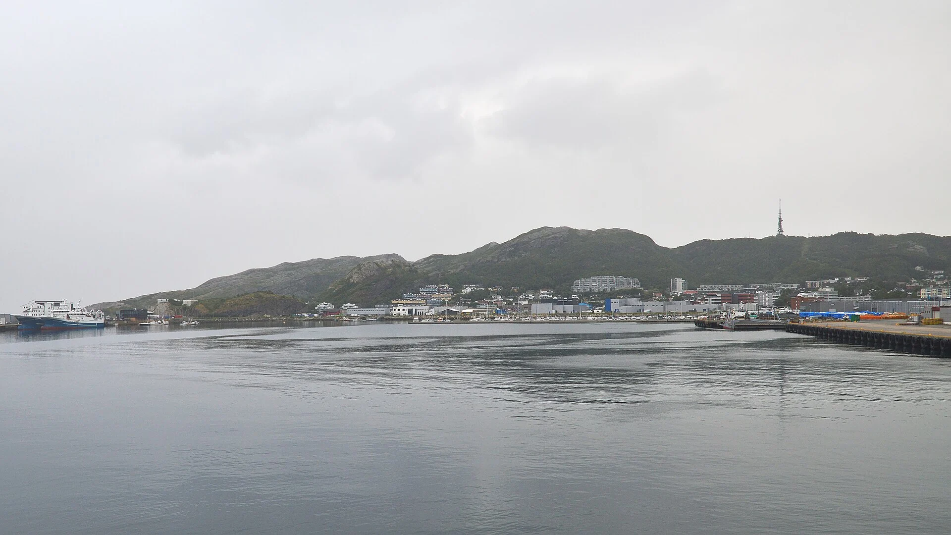Blick vom Wasser auf den Hafen und die Stadt Bodø mit Fähre, Kaianlagen und dem Sendemast auf dem Rønvikfjellet.