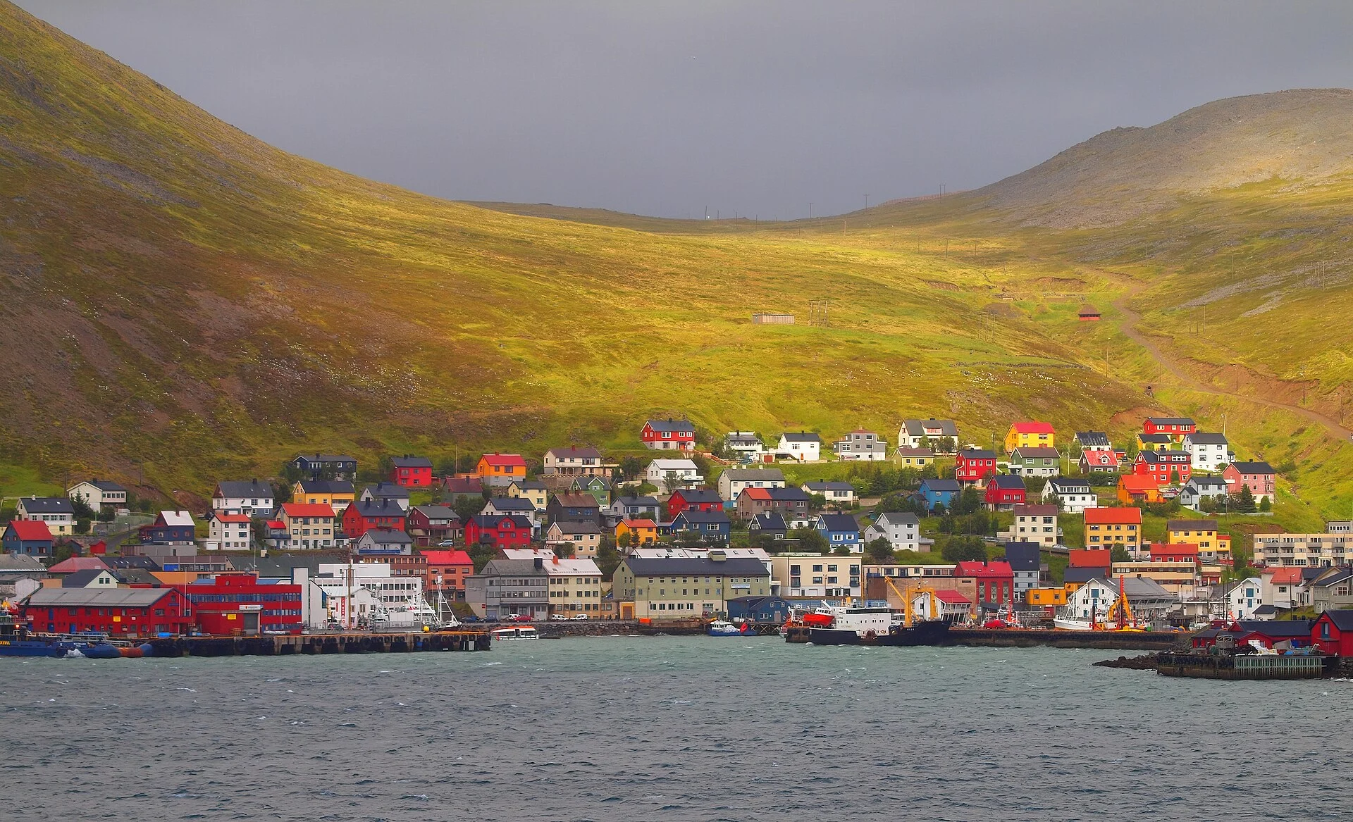 Blick vom Wasser auf den Ort Honningsvåg mit bunten Holzhäusern und Hafenanlagen am Fuß kahler Hügel.