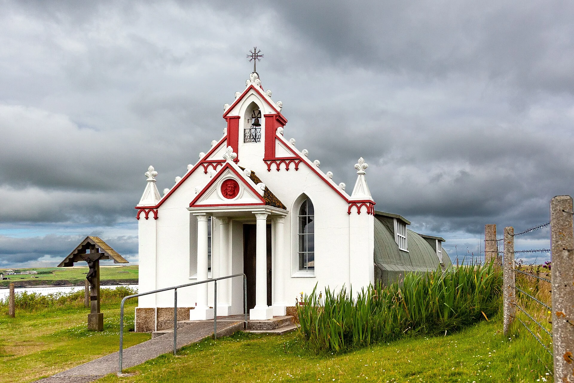 Die weiß-rote Fassade der Italian Chapel auf Lamb Holm bei Kirkwall, dahinter die Wellblechhütte, unter bewölktem Himmel.