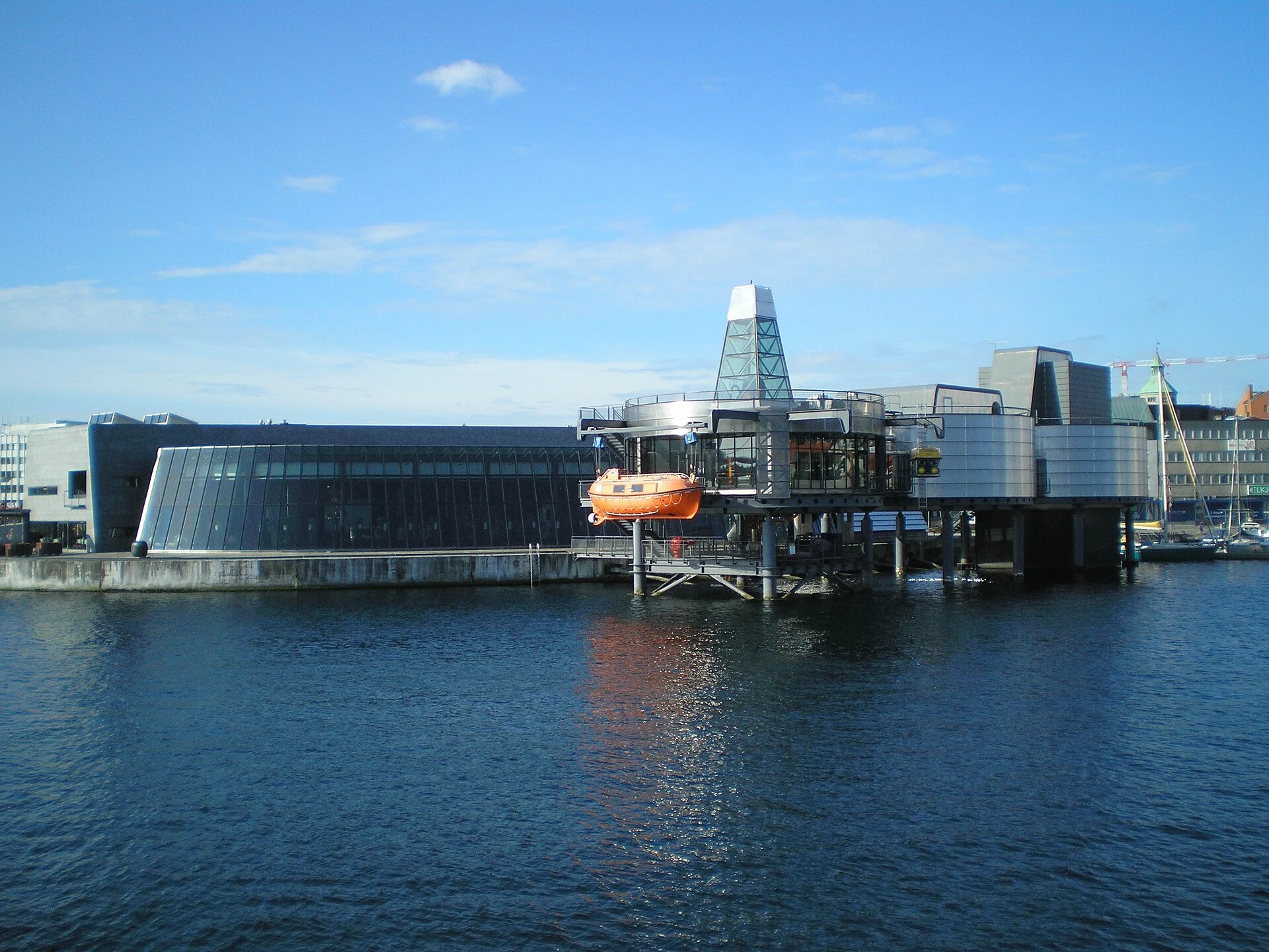 Das Norwegische Erdölmuseum in Stavanger vom Hafenbecken aus, mit Rettungsboot und Bohrturm-Nachbau über dem Wasser.