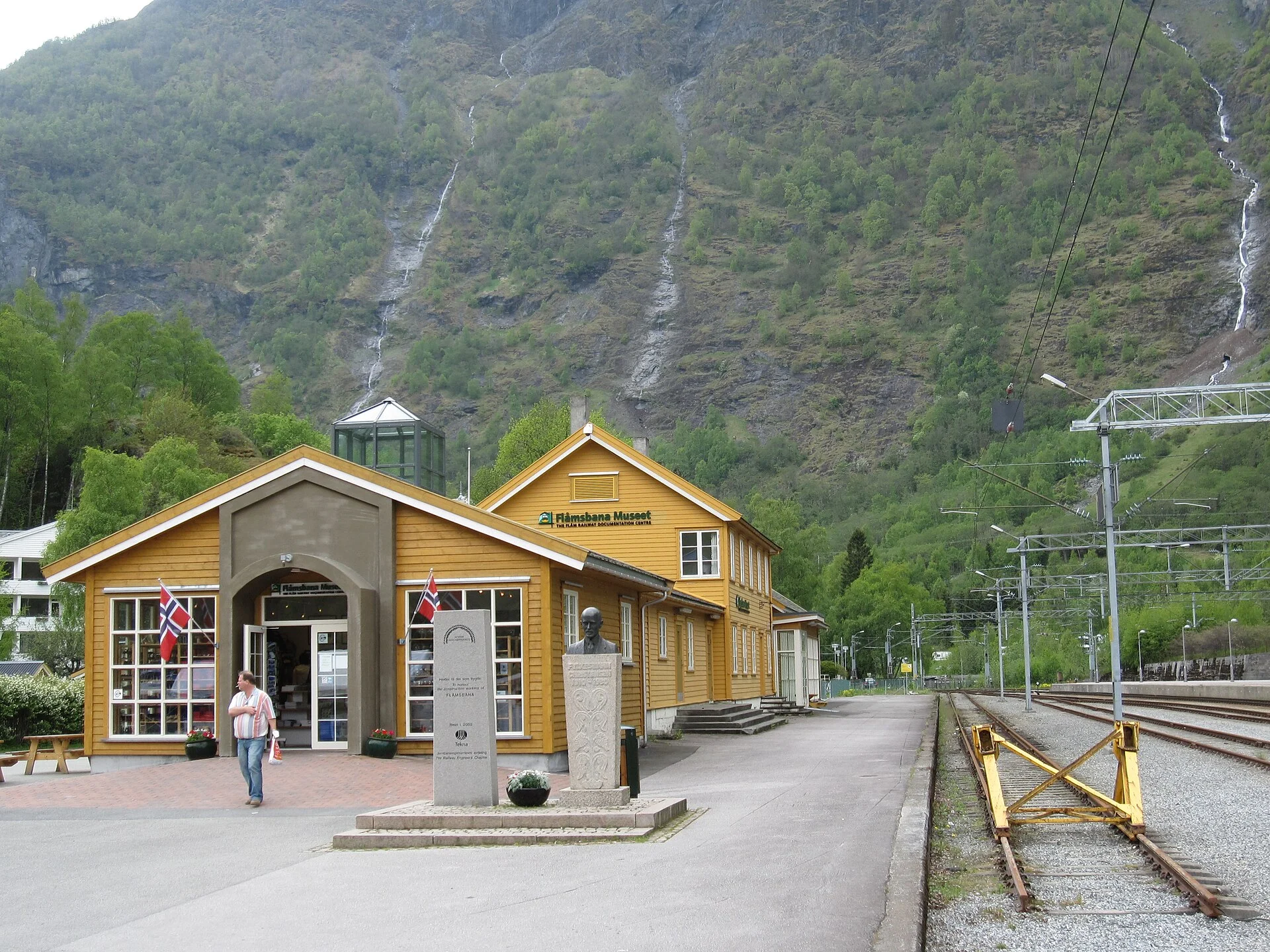 Das gelbe Holzgebäude des Flåmsbana Museums im alten Bahnhof von Flåm mit Schild, Denkmal und Bahngleisen.