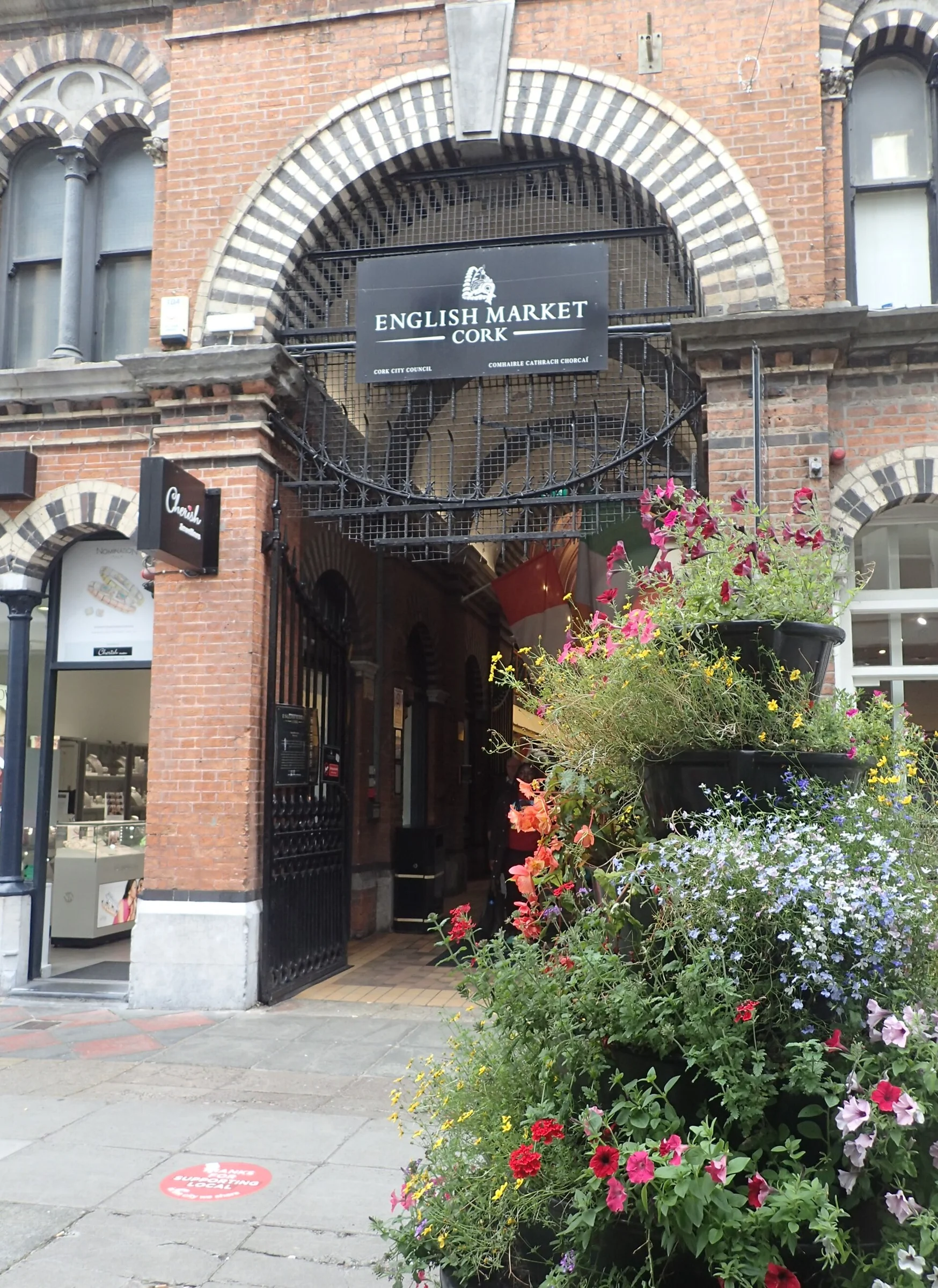 Torbogen mit Schild „English Market Cork“ und Blumenkübeln am Eingang des English Market in Cork.