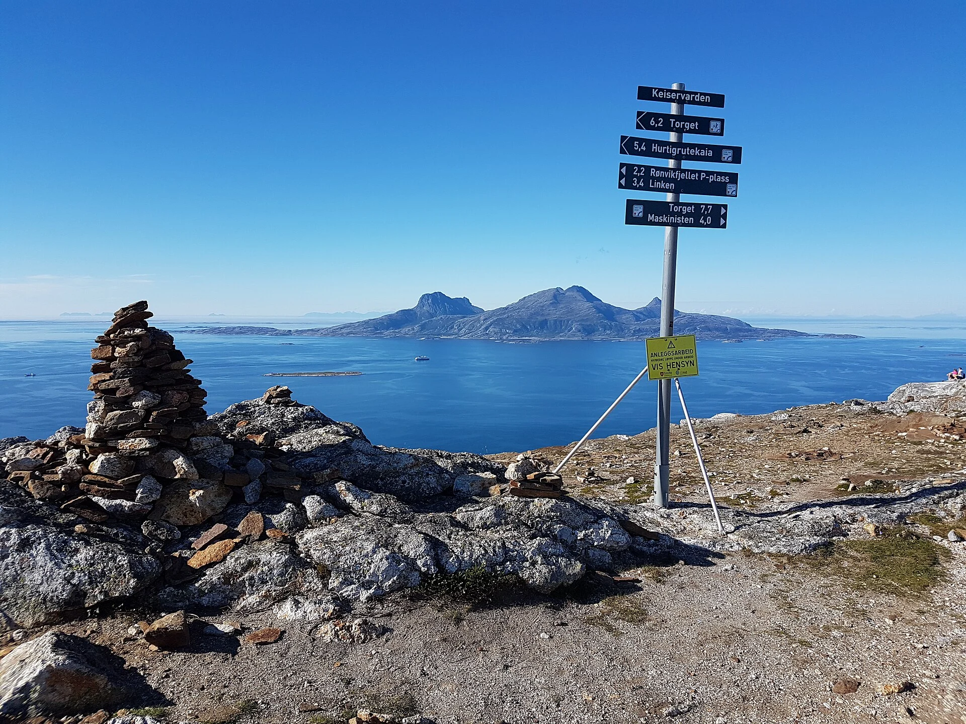 Wegweiser mit der Aufschrift „Keiservarden“ und Steinvarde auf dem Gipfelplateau des Keiservarden bei Bodø, dahinter der Fjord und die Insel Landegode.