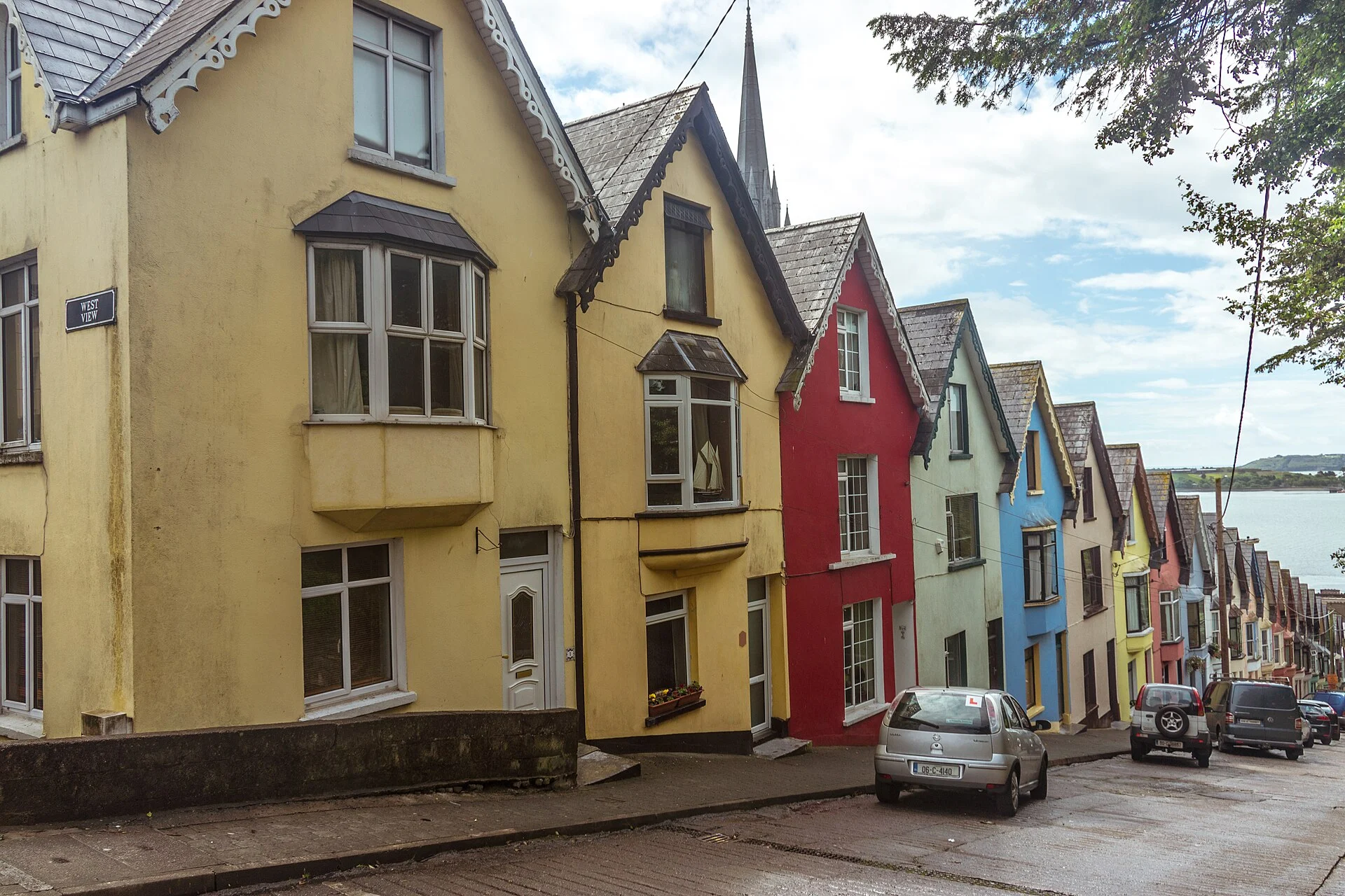 Die bunte Häuserzeile West View in Cobh mit Straßenschild, dahinter der Turm der St.-Colman-Kathedrale und das Hafenbecken.