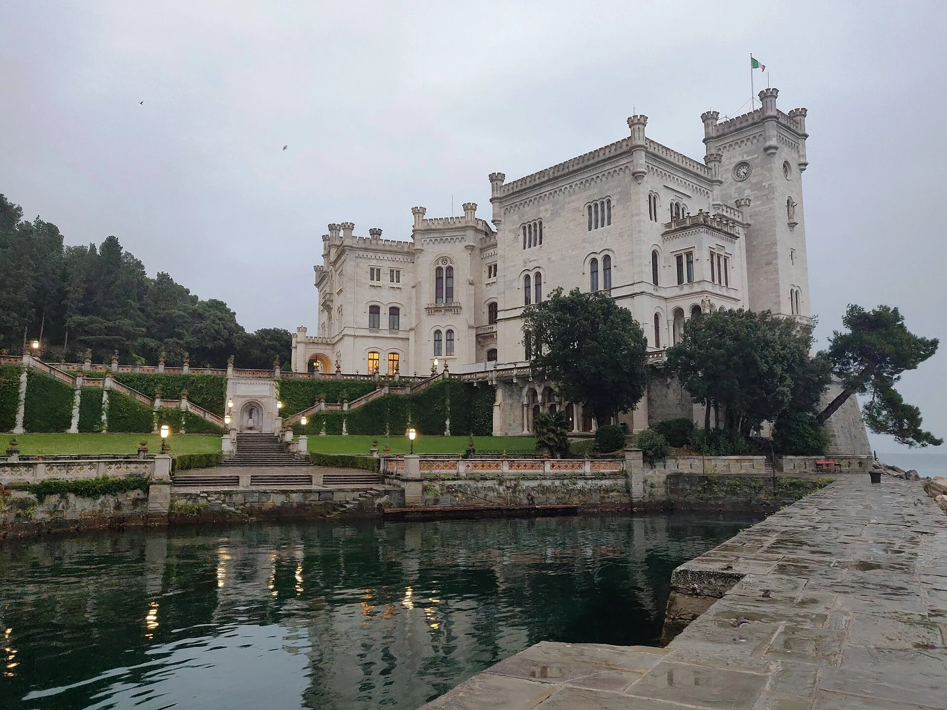 Schloss Miramare in Triest vom Hafenbecken aus mit Terrassen, Treppen und beleuchteten Fenstern in der Dämmerung.