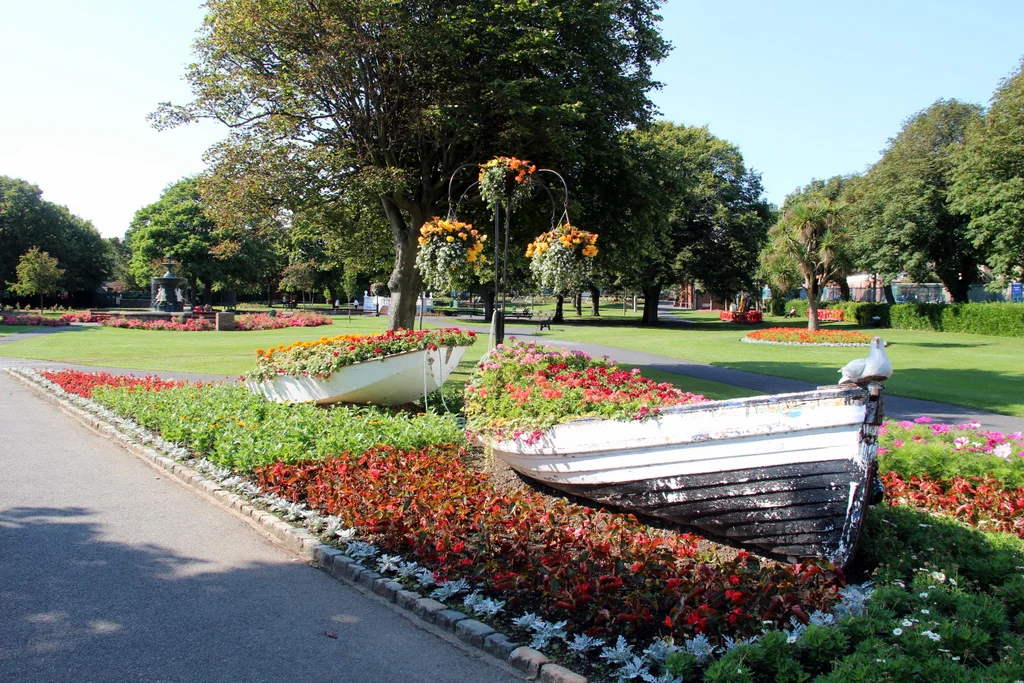 Blumenbeete mit bepflanzten Ruderbooten und Hängeampeln im People's Park in Dún Laoghaire, im Hintergrund Rasenflächen und alter Baumbestand.