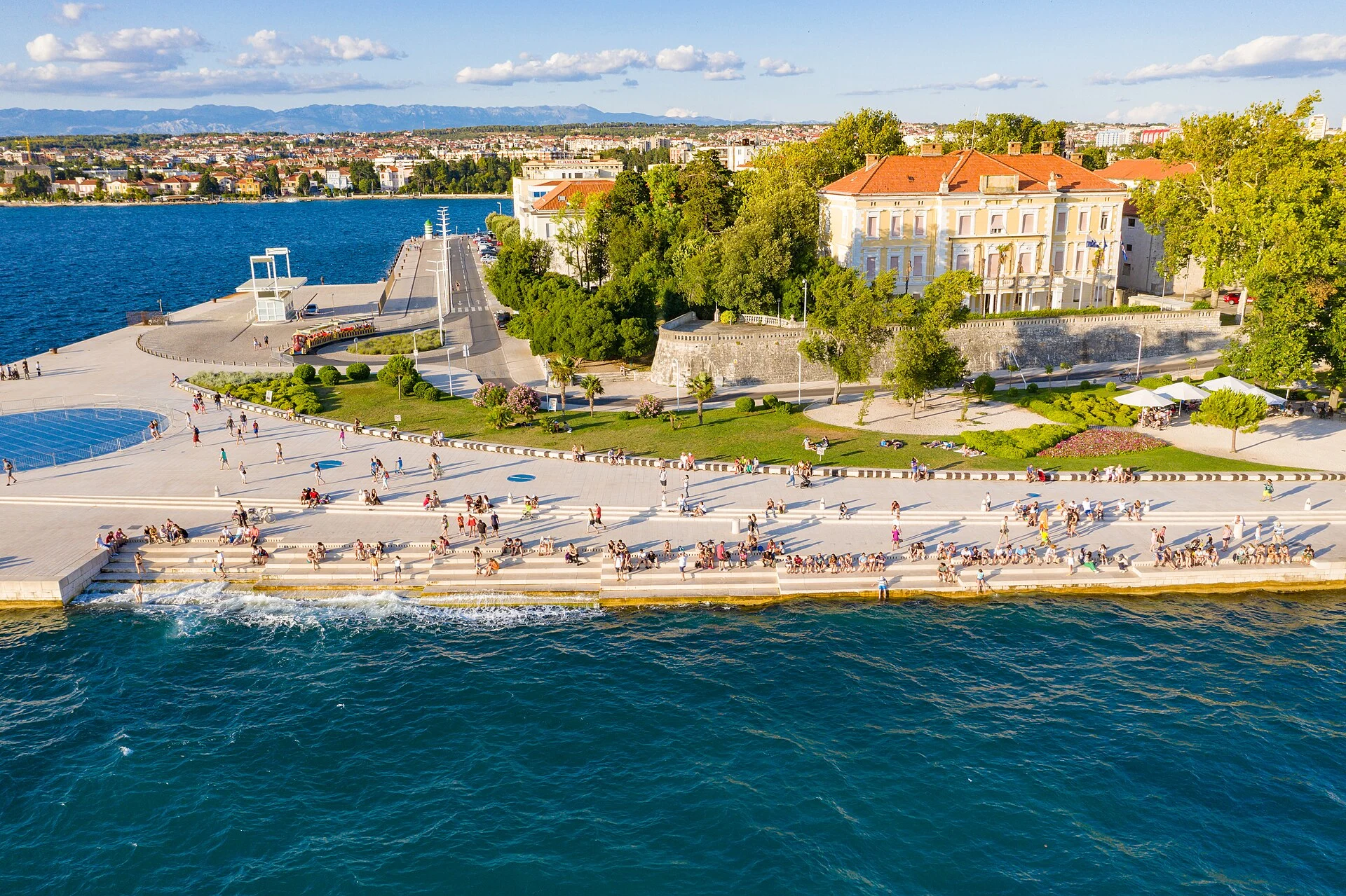 Luftbild der Meeresorgel in Zadar mit den Uferstufen, der Promenade und den Gebäuden der Halbinsel im Hintergrund.