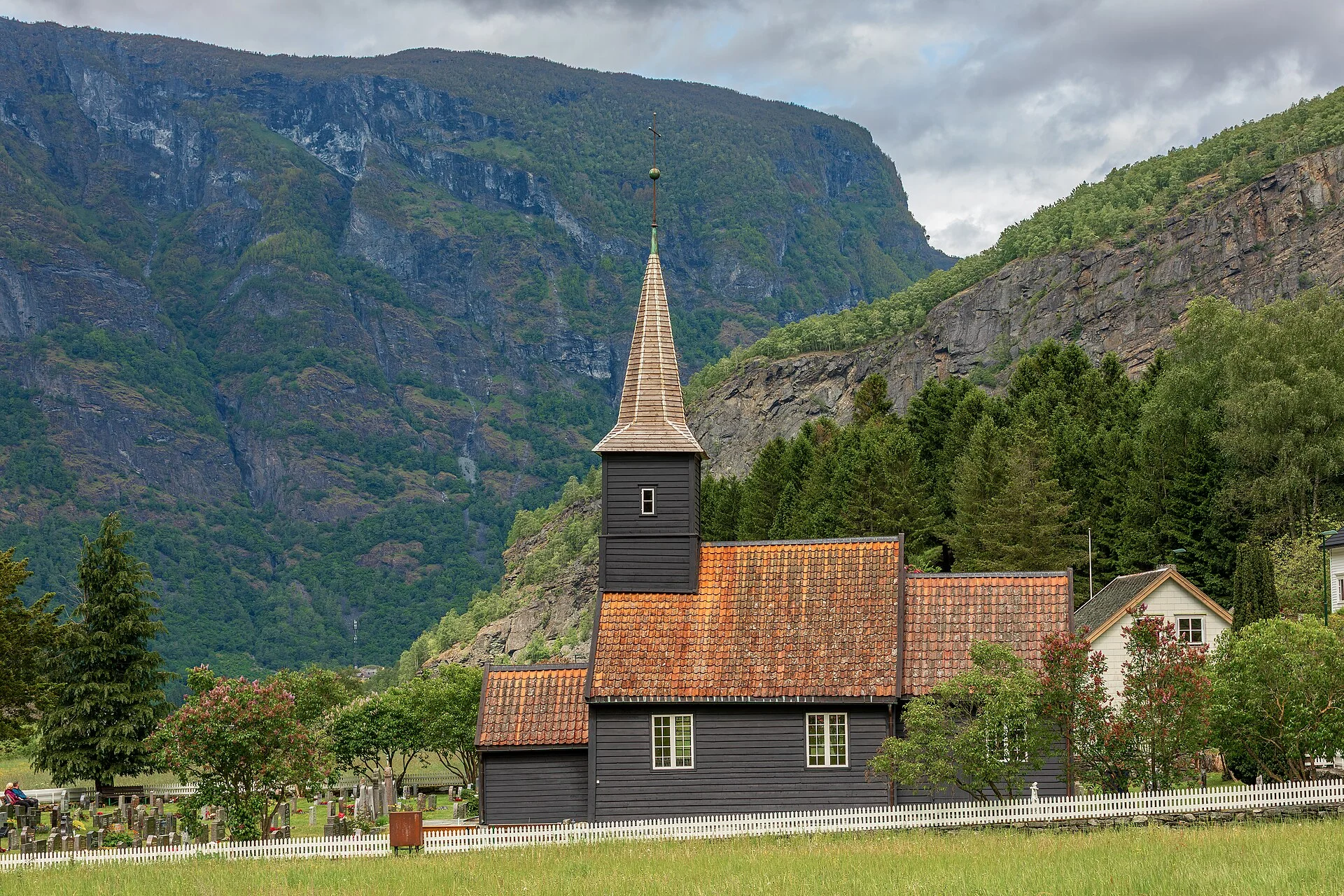 Die schwarze Holzkirche Flåm kyrkje mit Dachreiter und Friedhof vor den steilen Felswänden des Flåmstals.