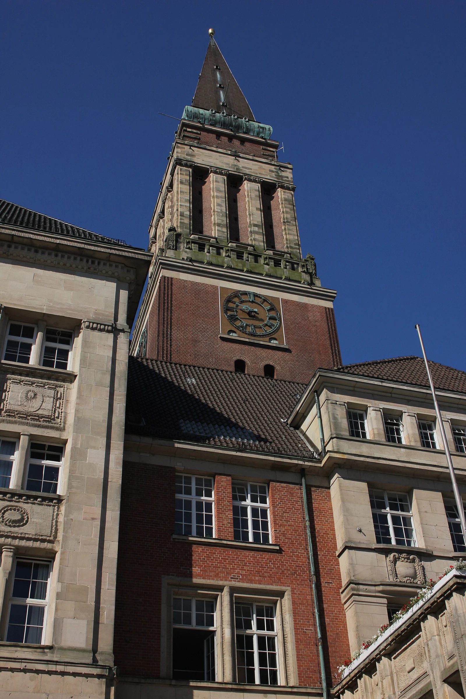 Der Backsteinturm des Kieler Rathauses mit Turmuhr, aufgenommen von unten gegen blauen Himmel.