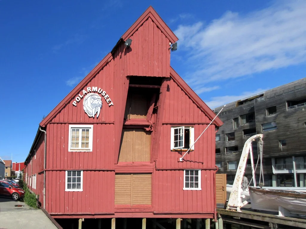 Das Polarmuseum in Tromsø, ein rotes Holzspeicherhaus von 1837 auf Pfählen am Wasser, bei Sonnenschein.
