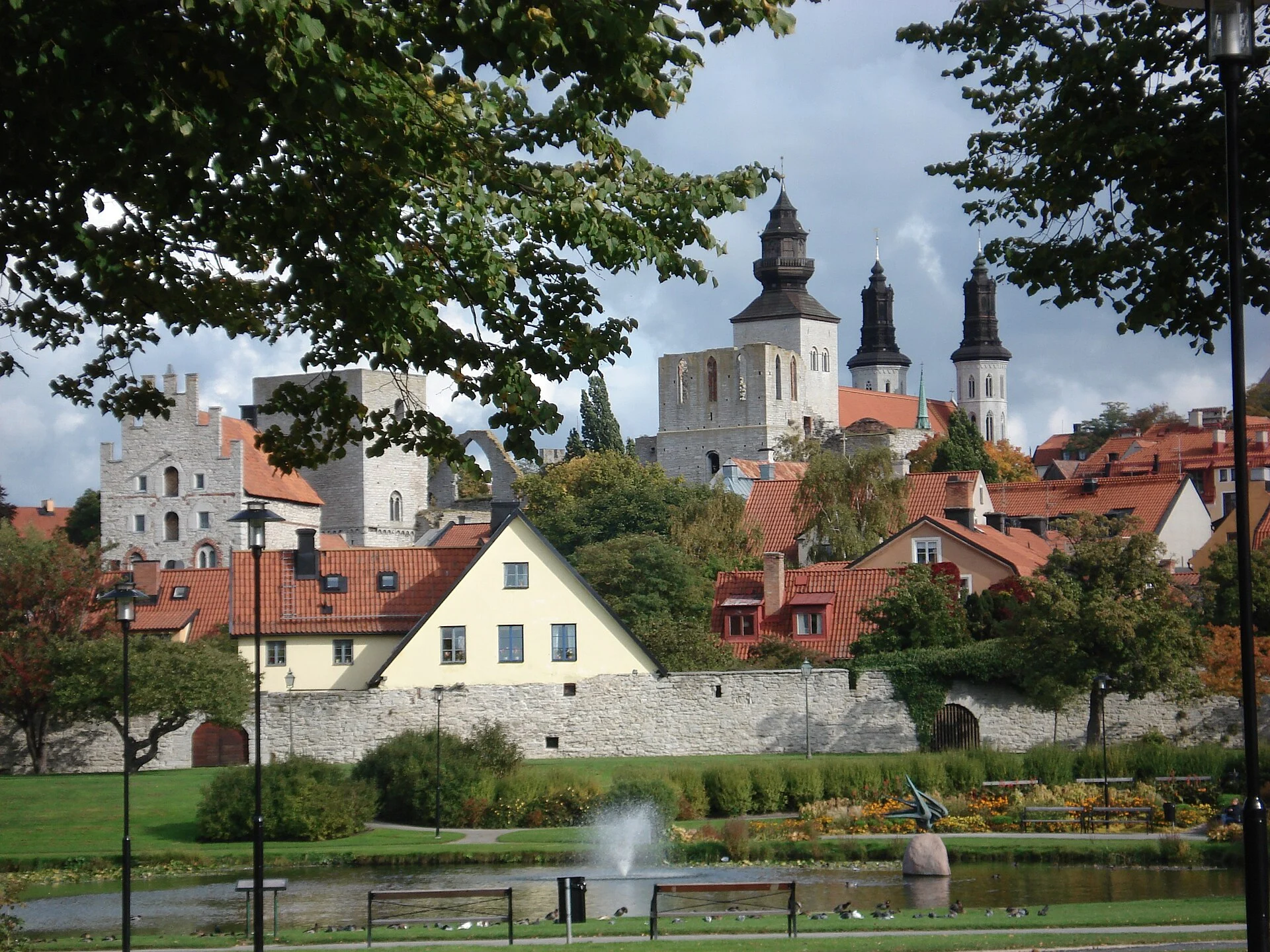 Der Park Almedalen in Visby mit Teich und Springbrunnen, dahinter Stadtmauer, Ruinen und die Türme des Doms.