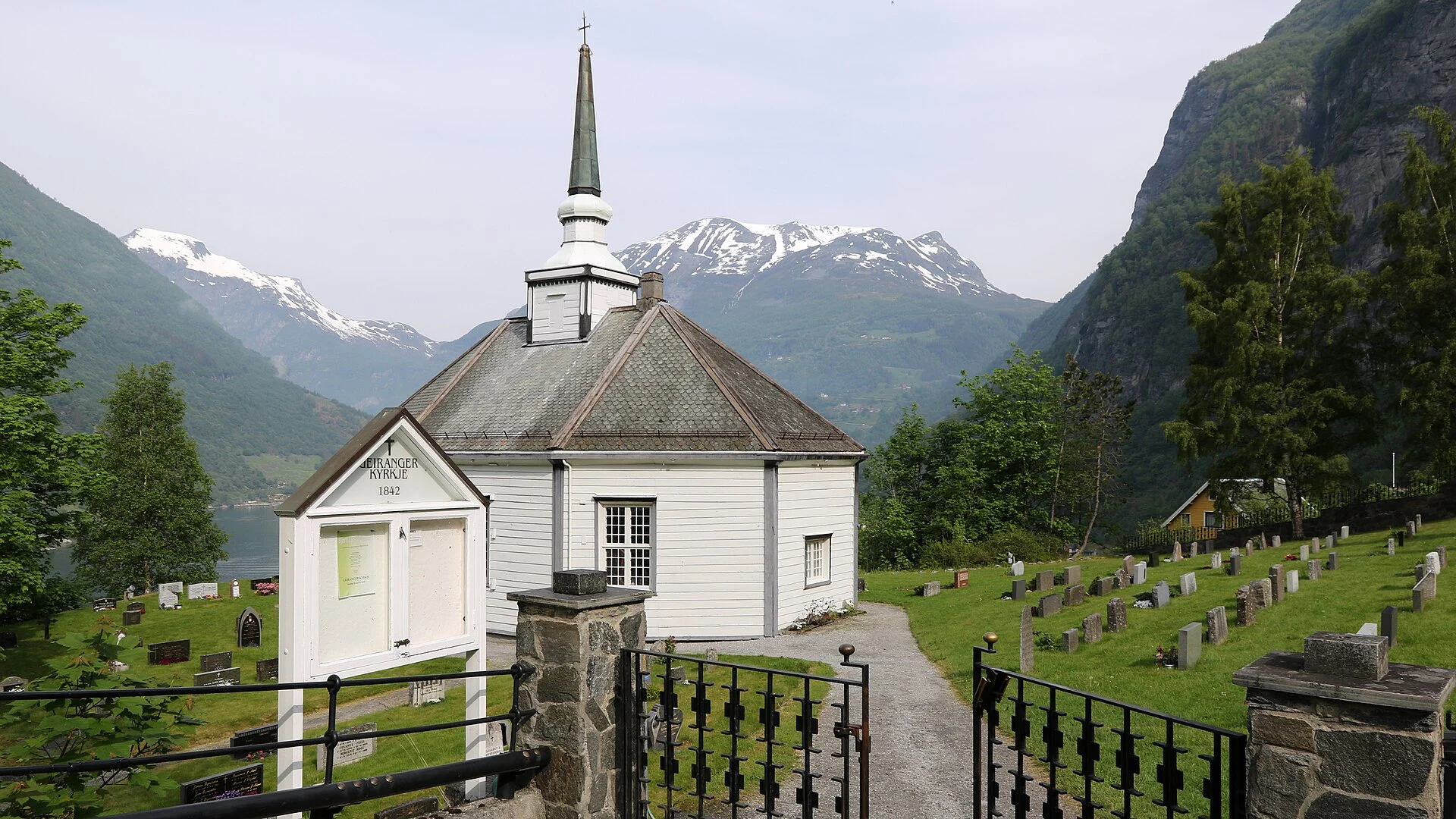 Eingangstor zum Friedhof von Geiranger kyrkje mit Schild „Geiranger Kyrkje 1842“, der weißen Oktogonkirche und den schneebedeckten Bergen über dem Fjord.