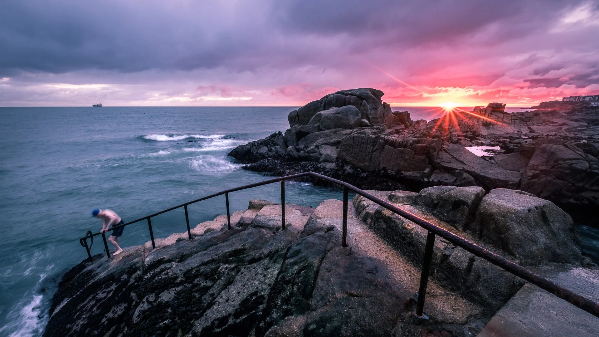 Sonnenaufgang am Forty Foot in Sandycove/Dún Laoghaire: Felsküste mit Betonstufen, Geländer und einem Schwimmer am Wasser.