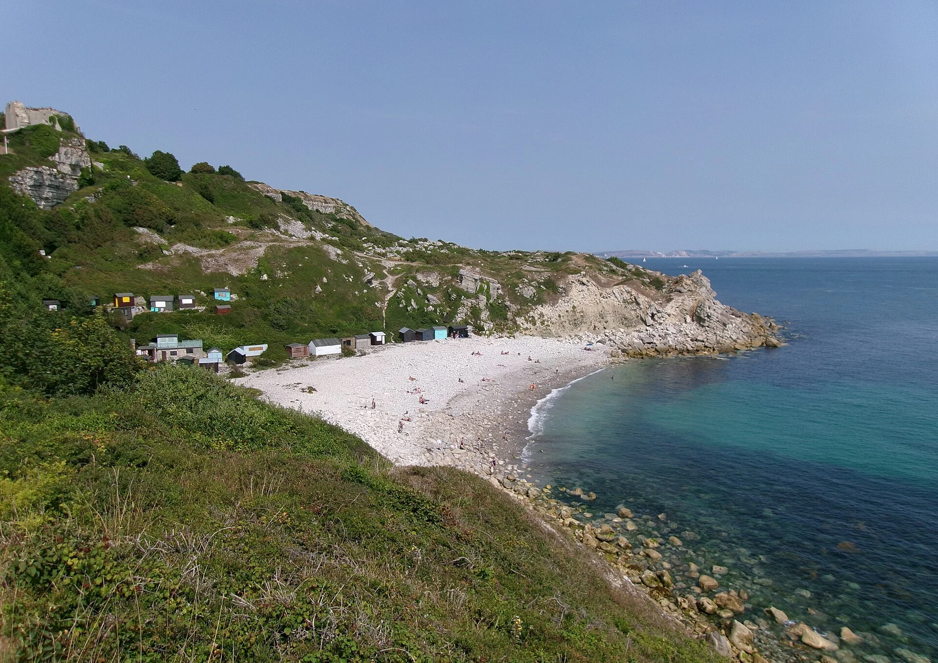 Church Ope Cove auf der Isle of Portland mit hellem Kiesstrand, bunten Strandhütten und der Ruine von Rufus Castle oben links.