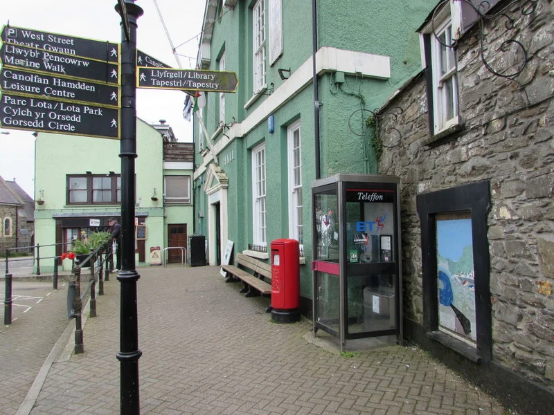 Zweisprachiger Wegweiser, roter Briefkasten und Telefonzelle vor der grün gestrichenen Town Hall mit Markthalle am Market Square in Fishguard.