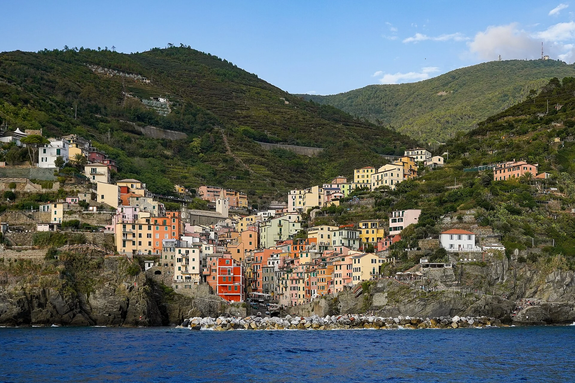 Blick vom Meer auf Riomaggiore in den Cinque Terre mit den bunt gestapelten Häusern über dem kleinen Hafen und den bewaldeten Hängen dahinter.