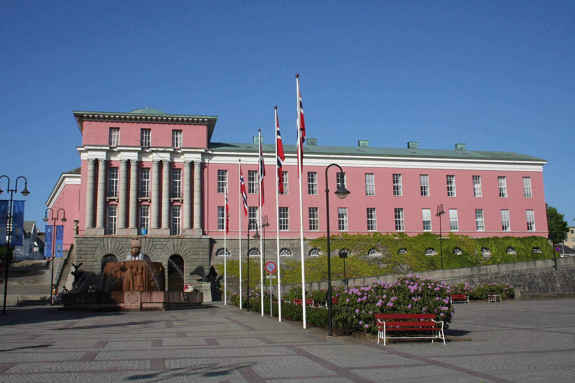 Das rosafarbene Rathaus (Rådhuset) von Haugesund mit norwegischen Flaggen und dem gepflasterten Platz des Rådhusparken davor.