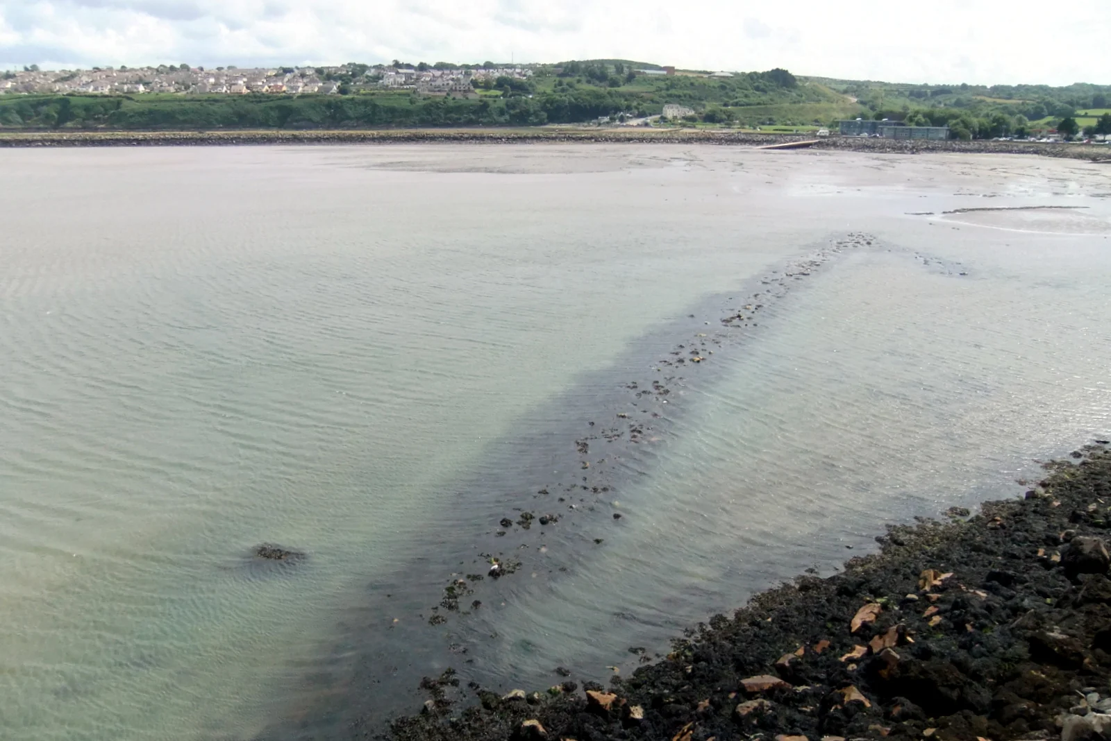 Weite Sandfläche der Bucht bei Goodwick mit alter Fischfalle im flachen Wasser und den Häusern von Goodwick am Hang.