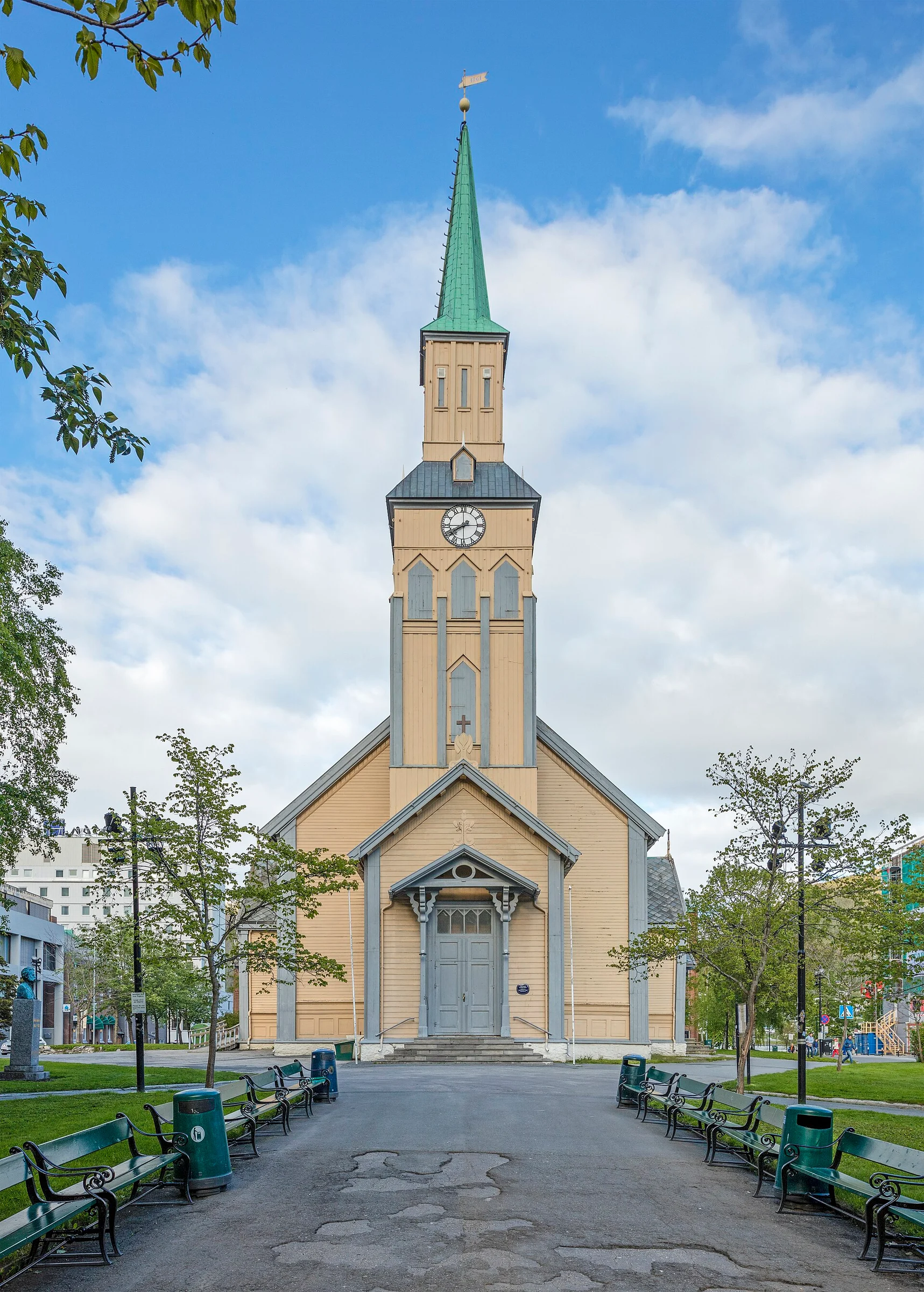 Frontansicht der gelben Holz-Domkirche von Tromsø mit grünem Turmhelm und Parkbänken davor.