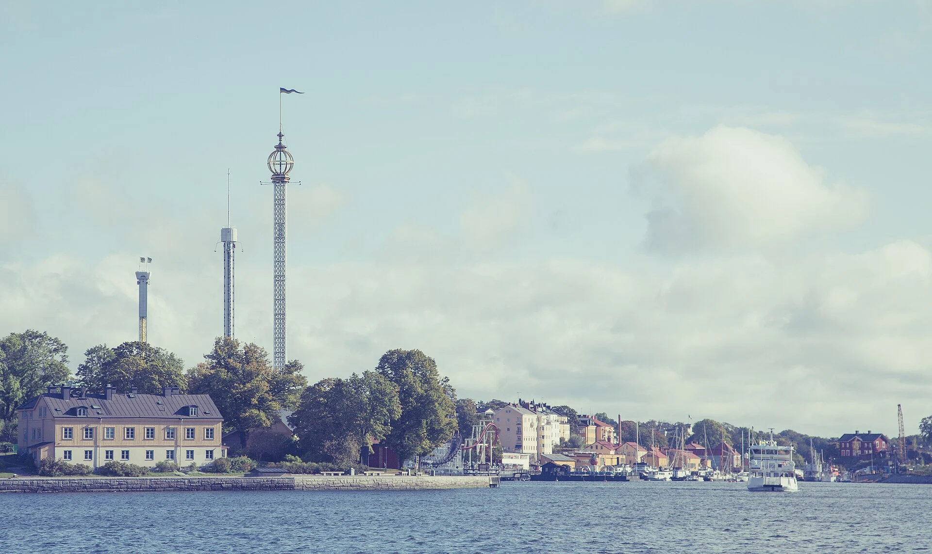 Blick vom Wasser auf die Insel Djurgården in Stockholm mit den Fahrgeschäftstürmen des Vergnügungsparks Gröna Lund und gelben Holzhäusern am Ufer.