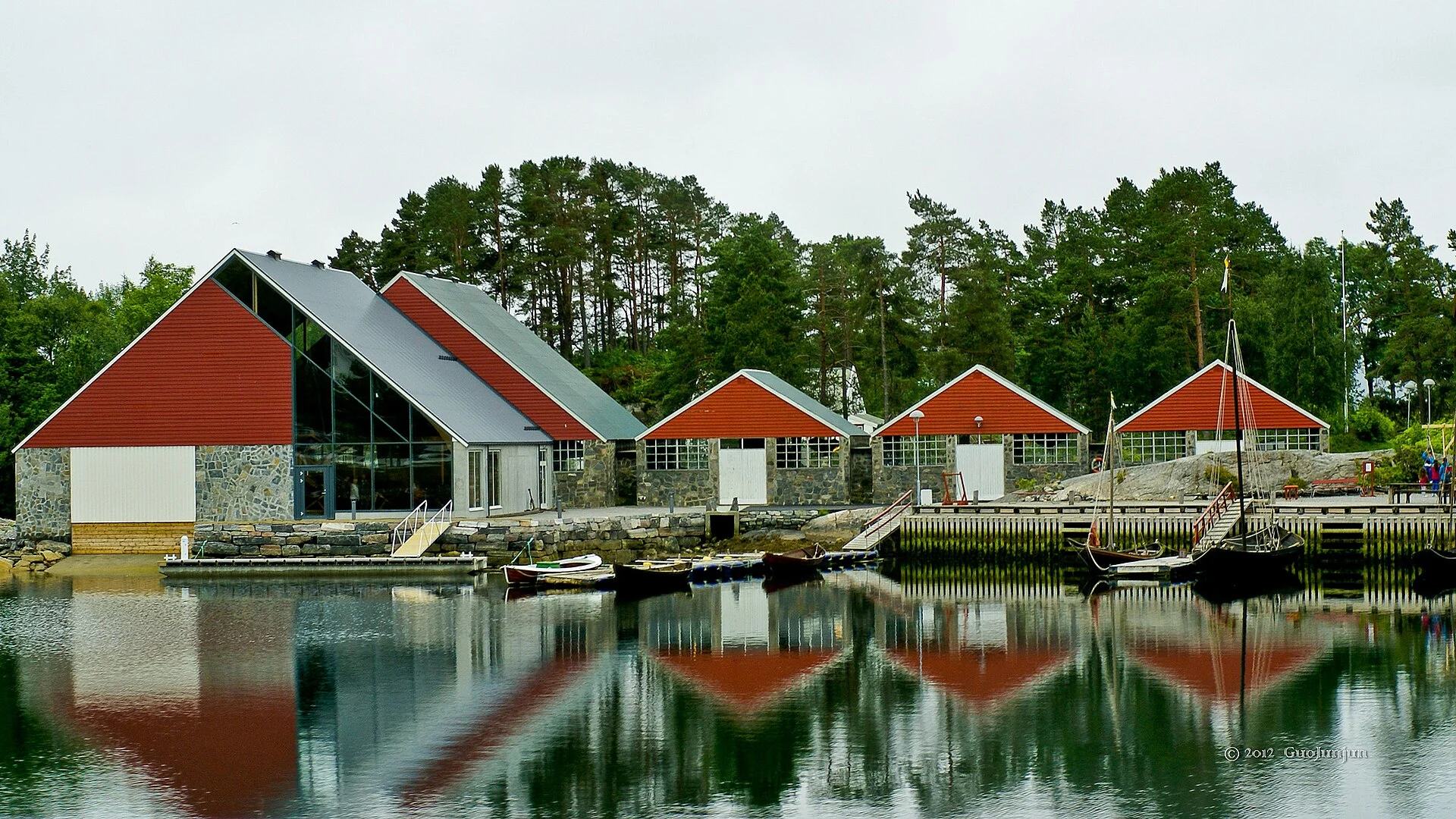 Rot gedeckte Museums- und Bootshallen des Sunnmøre Museums bei Ålesund spiegeln sich im ruhigen Wasser, rechts liegen offene Holzboote.