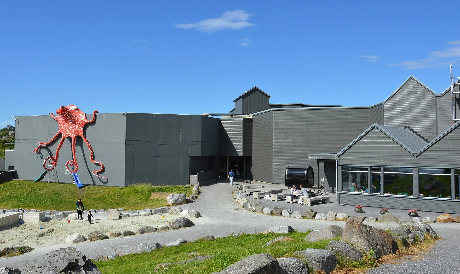 Graues Hauptgebäude des Atlanterhavsparken in Ålesund mit rotem Kraken-Spielgerät an der Fassade und Außenanlage.