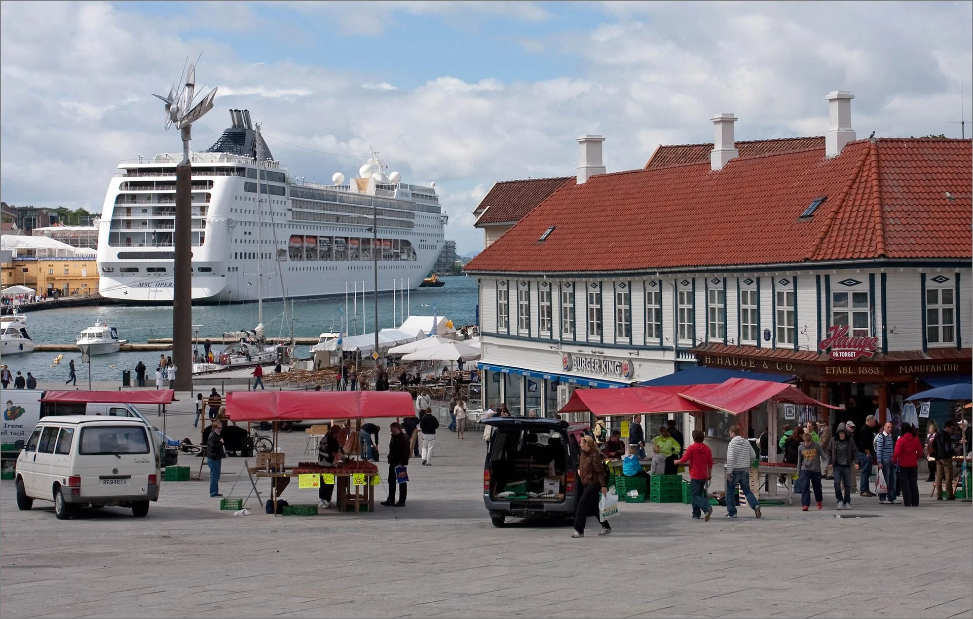 Marktstände mit roten Planen auf dem Torget in Stavanger, dahinter das Fischmarkt-Gebäude und ein Kreuzfahrtschiff im Hafen.