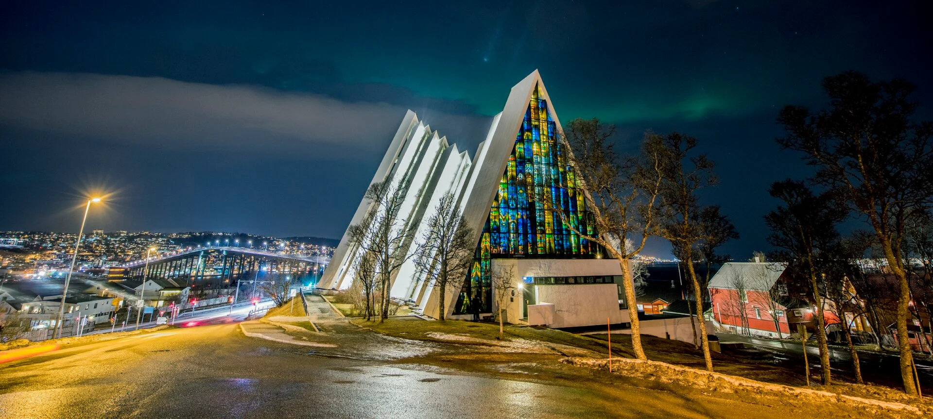 Die Eismeerkathedrale in Tromsø bei Nacht mit beleuchtetem Glasmosaik der Ostwand und Nordlicht am Himmel.