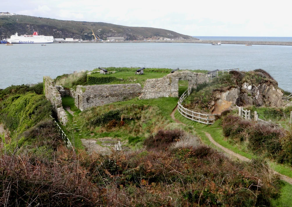 Blick von oben auf die Ruinen und Kanonen von Fishguard Fort auf Castle Point, dahinter der Hafen von Fishguard mit Fähre.