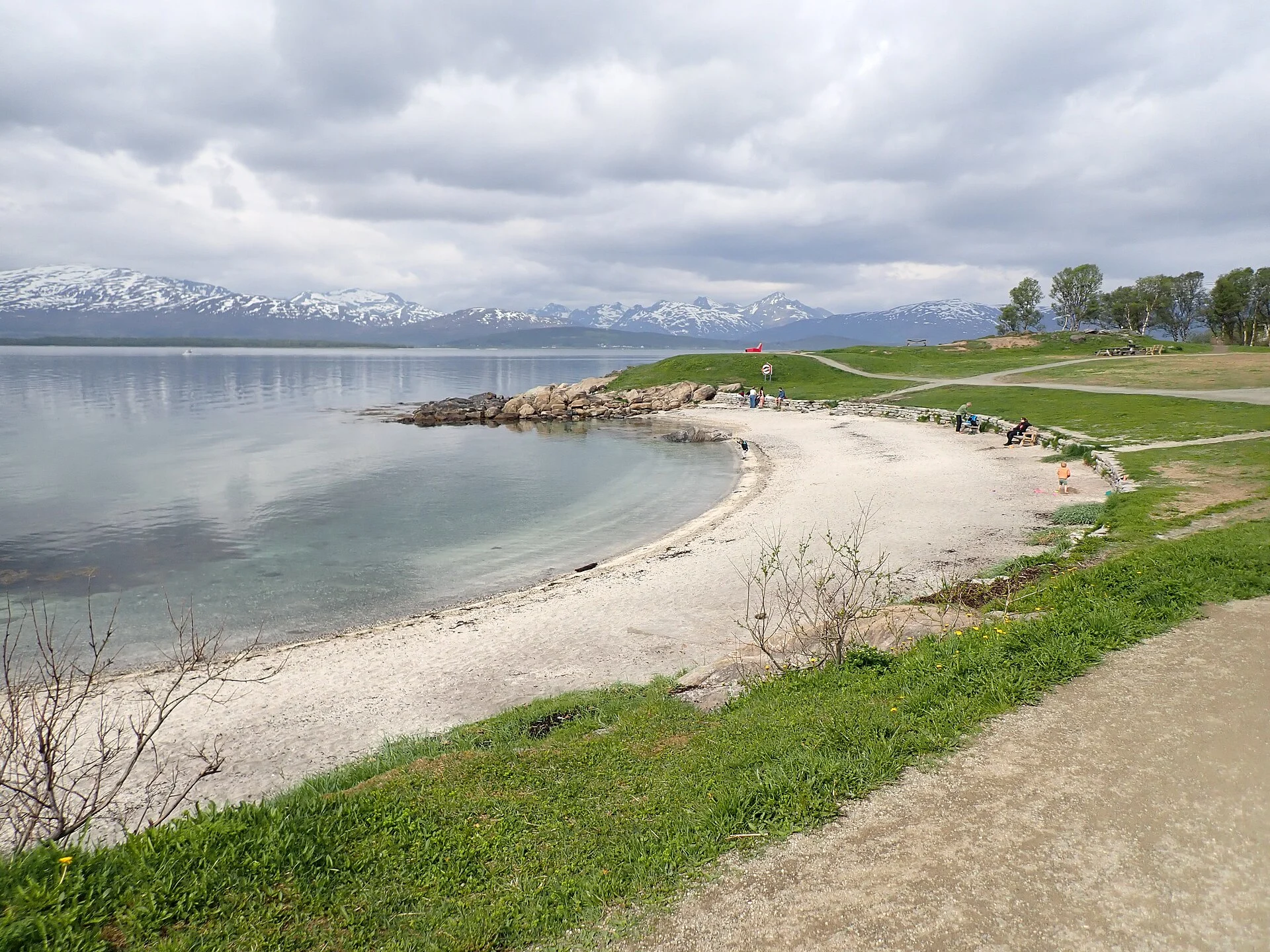 Heller Kies- und Sandstrand der Telegrafbukta an der Südwestseite von Tromsøya mit Wiese, Wegen und schneebedeckten Bergen der Kvaløya.