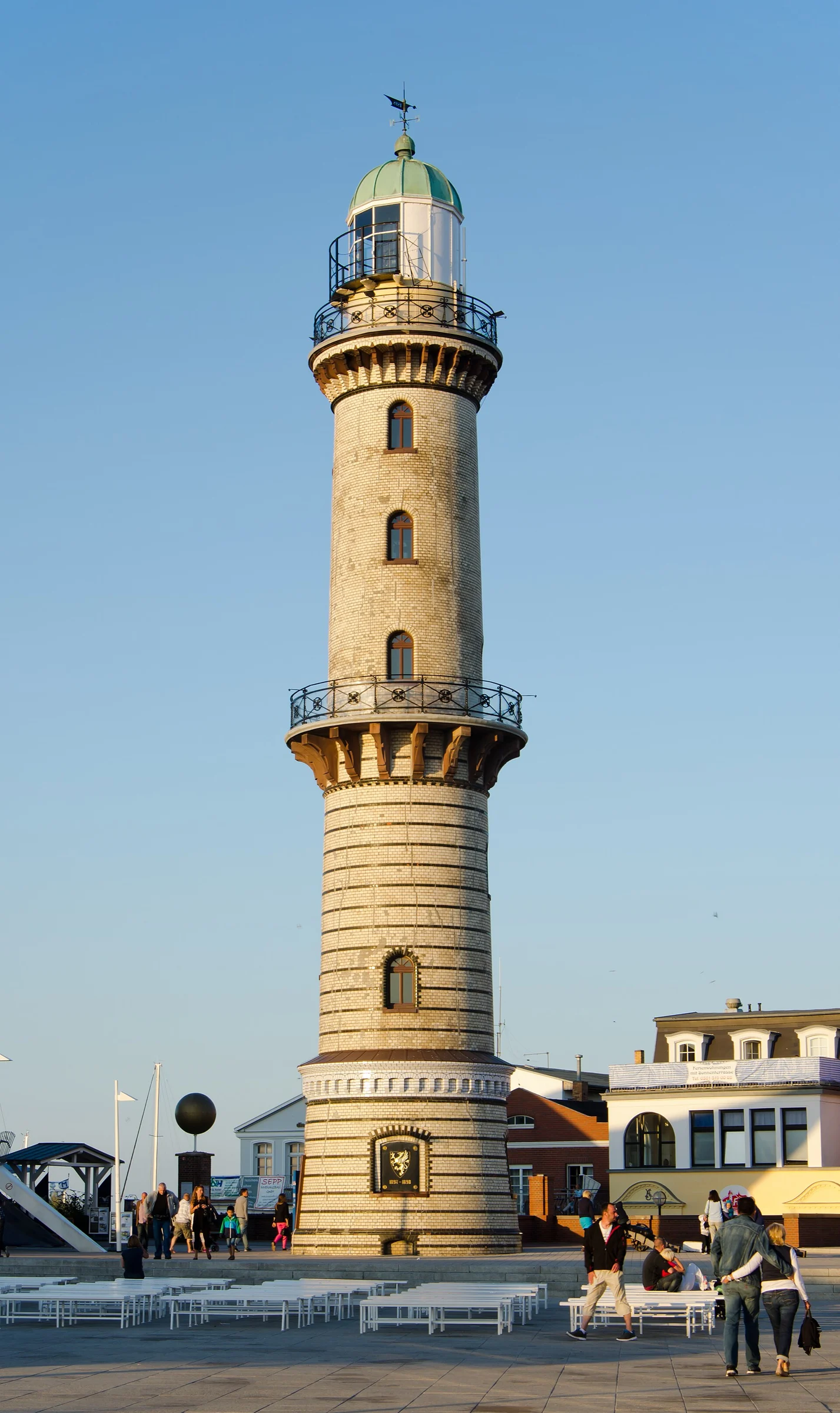 Der 37 Meter hohe Leuchtturm Warnemünde am Strandübergang im Abendlicht, davor der gepflasterte Platz.