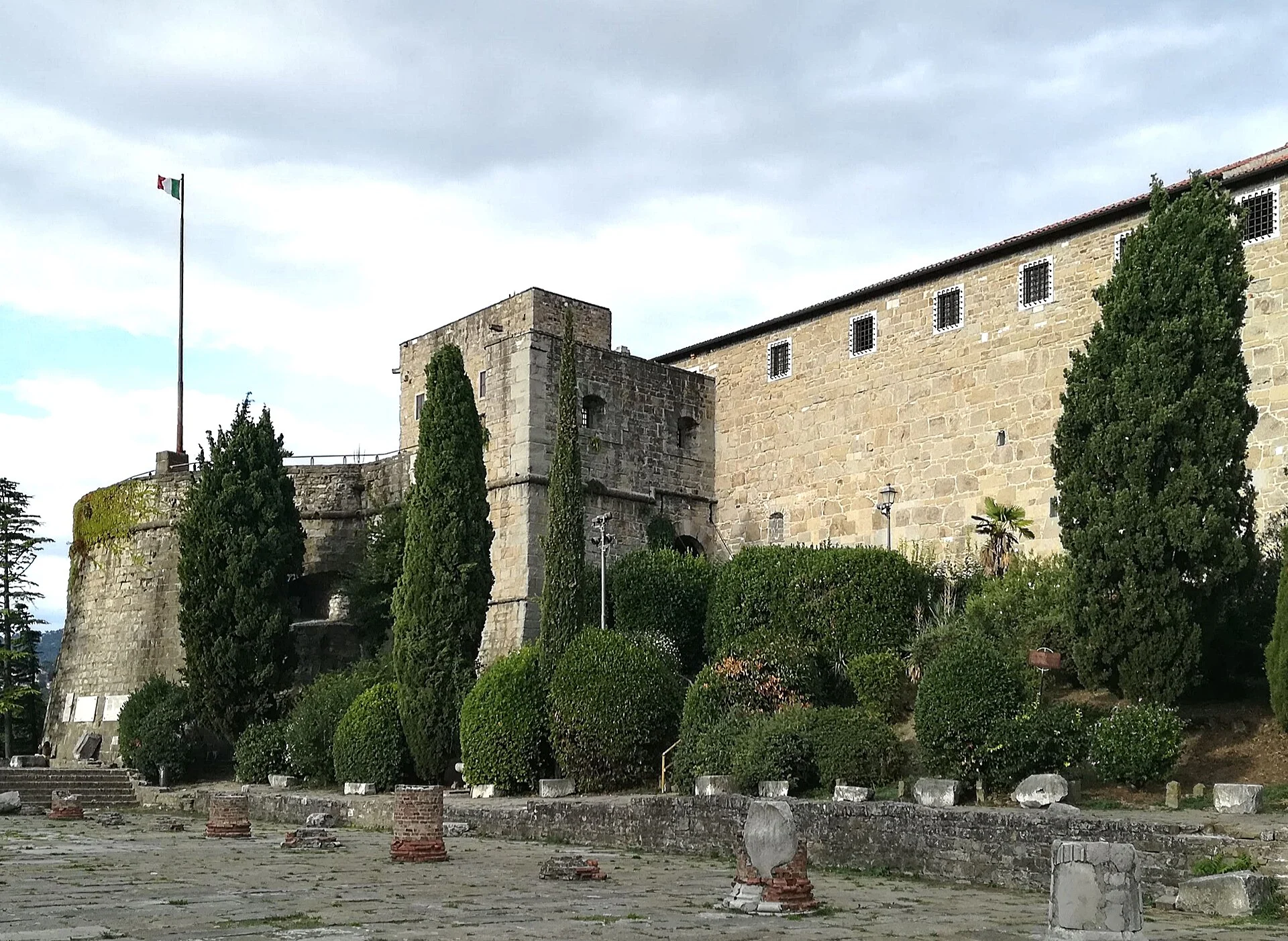 Das Castello di San Giusto in Triest mit runder Bastion, italienischer Flagge und den Ruinen des römischen Forums im Vordergrund.