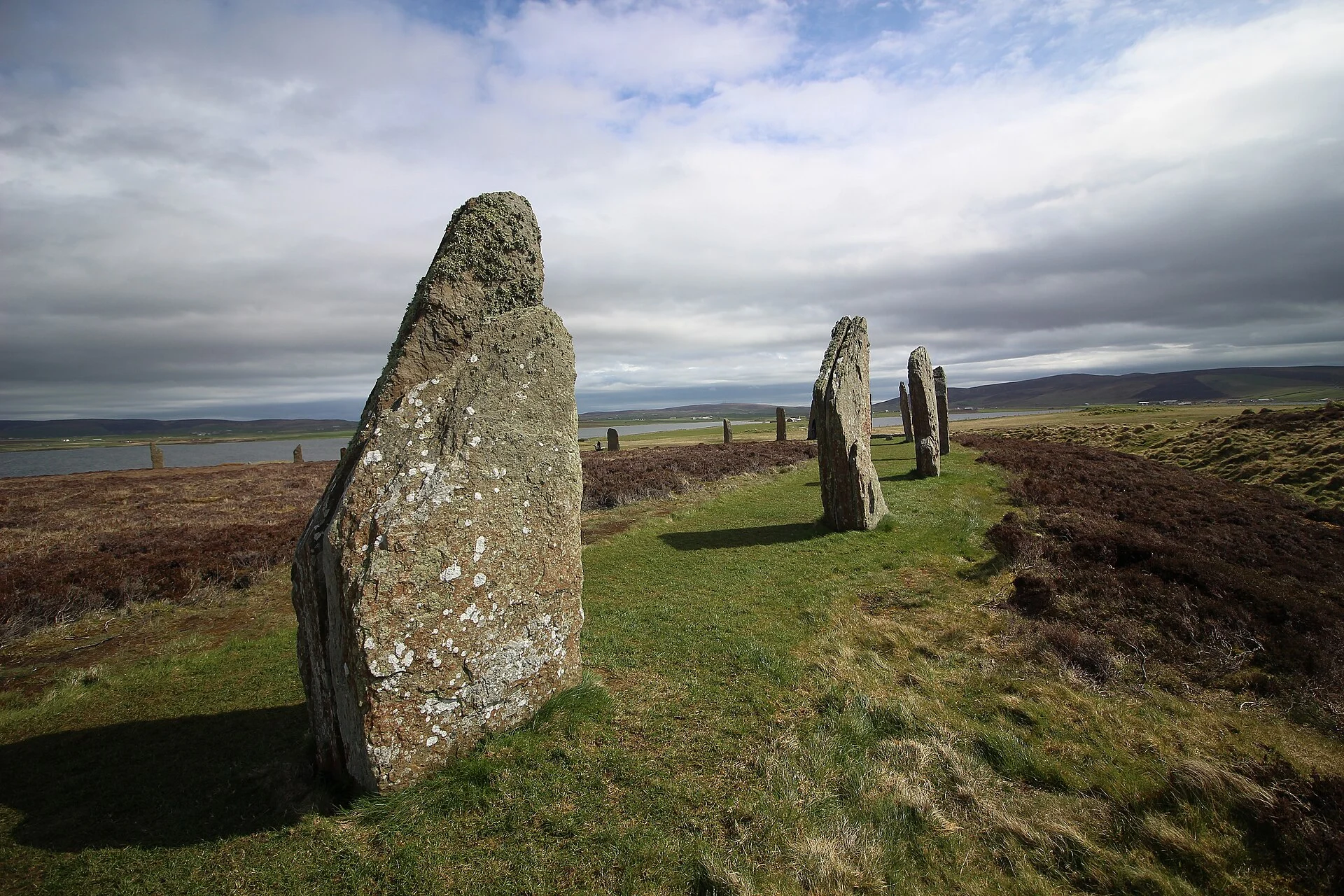 Reihe hoher Steine des Ring of Brodgar auf Orkney entlang des Grabens, im Hintergrund der See und sanfte Hügel.