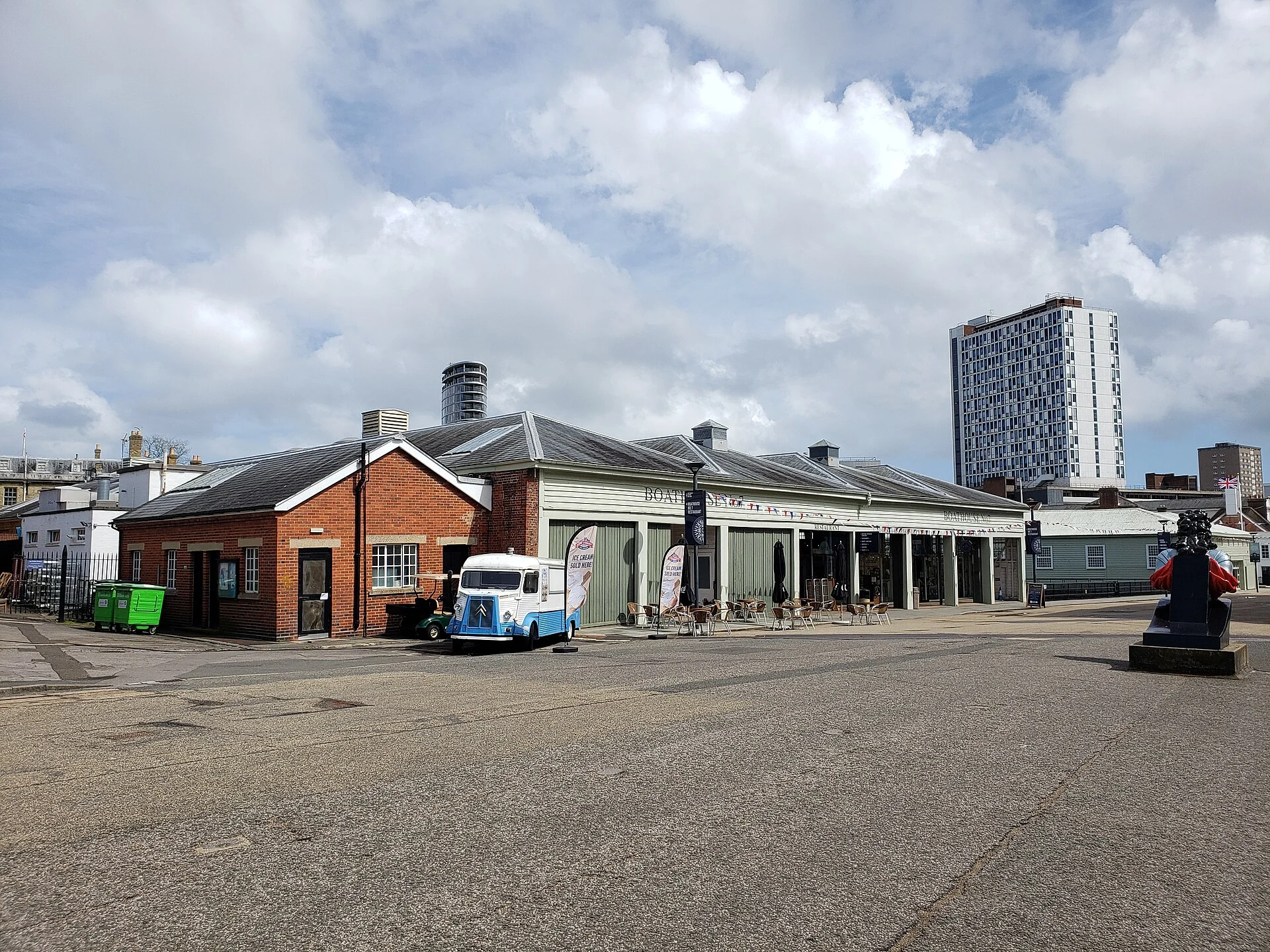 Boathouse No 7 mit Café-Terrasse auf dem Gelände des Portsmouth Historic Dockyard, im Hintergrund ein Hochhaus.