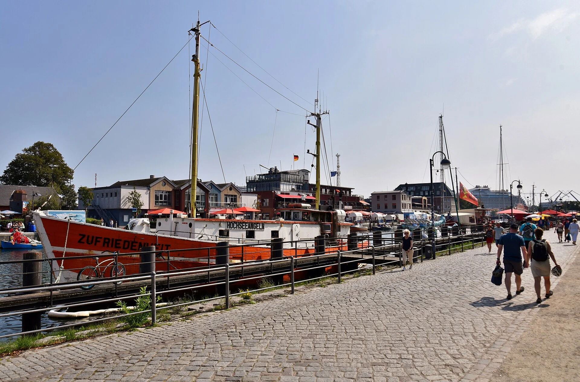 Der Alte Strom in Warnemünde mit festgemachtem rotem Kutter „Zufriedenheit“, Promenade mit Kopfsteinpflaster und Imbissbuden vor den Häusern am Westufer.